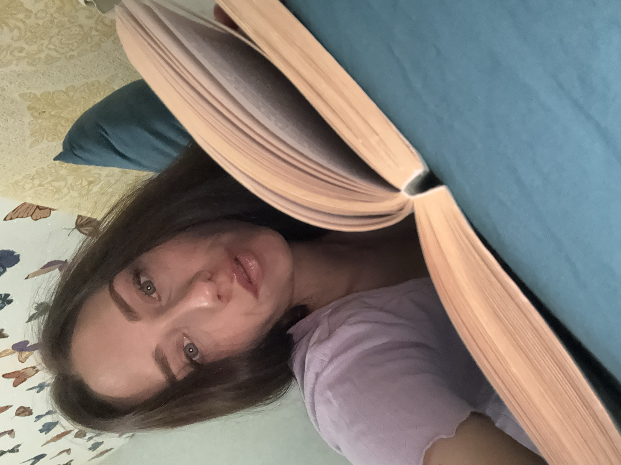 A person is visible in the image. The person has brown hair, fair skin, and their expression is neutral. They are partially obscured by an open book. The book has a light blue cover and off-white pages. The backdrop includes a floral patterned wall and a dark blue pillow. There is no information to determine if the image was taken in Saint Petersburg, Russia.