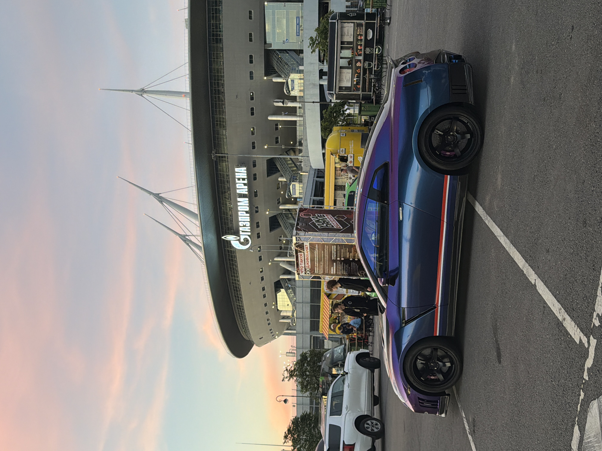 A Nissan GT-R with a holographic paint job is parked in a paved lot in front of the Gazprom Arena stadium at sunset. The sky is a gradient of pink, orange, and blue, with wispy clouds. Several food trucks are set up in the background, and a white SUV is parked to the left of the GT-R. Two young men are standing near a food truck, looking at a display. The stadium is large and modern, with distinctive white spires reaching towards the sky. The scene appears to be a public gathering or event.