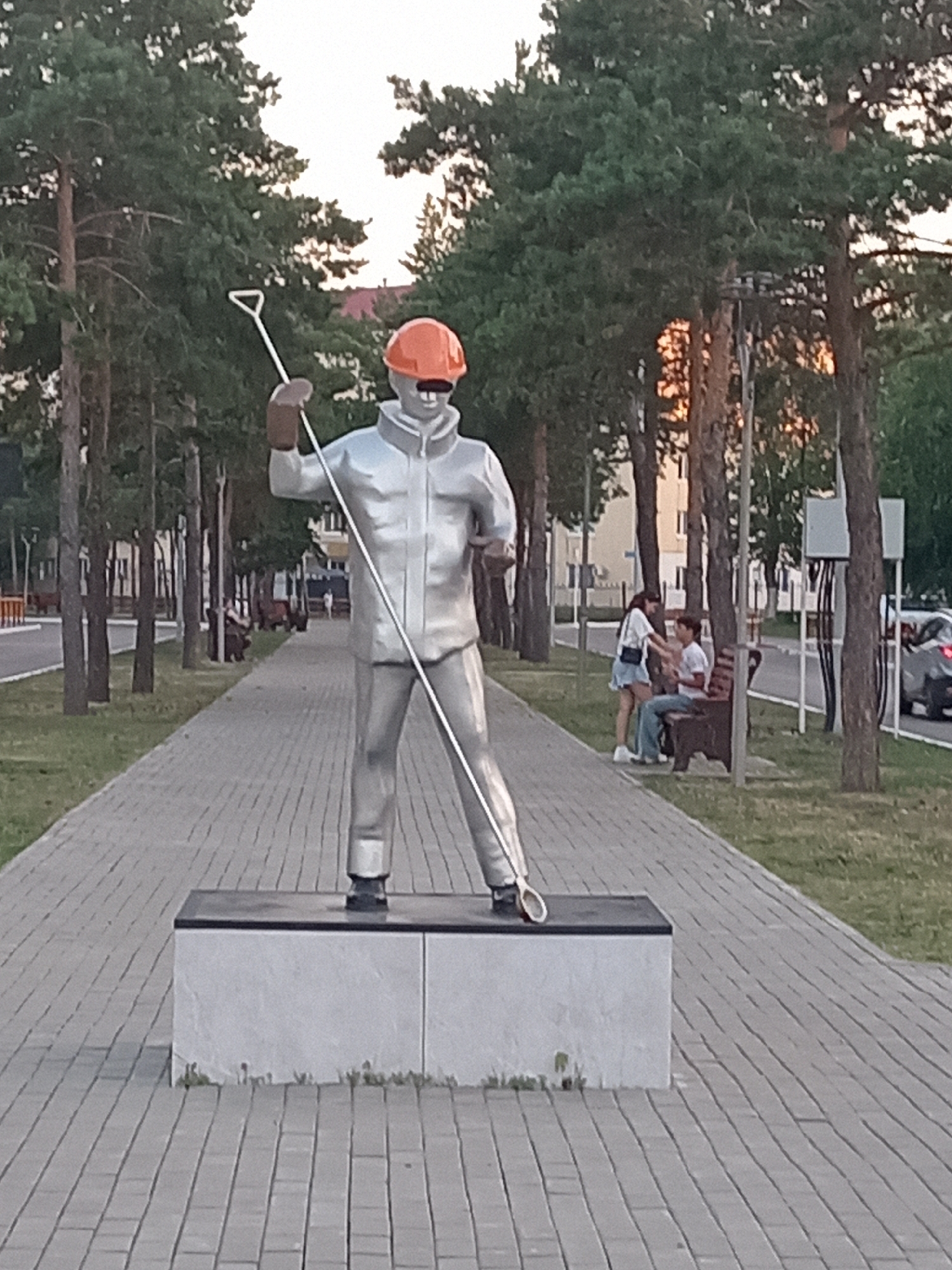 A life-sized metallic sculpture of a worker wearing an orange hard hat and holding a shovel stands on a grey stone base along a paved walkway lined with tall green trees. The sculpture is positioned centrally in the foreground, facing slightly to the left. Behind the sculpture, a couple is seated on a wooden bench, with the woman standing and interacting with the man who is seated. The walkway extends into the distance, bordered by grass and more trees. A paved road with passing cars is visible to the right of the walkway, beyond the bench and trees. The sky is overcast with soft light, suggesting it is either early morning or late evening. This sculpture is located in the city of Rudny, Kazakhstan, on Oktyabrskaya Street, a well-known landmark.
