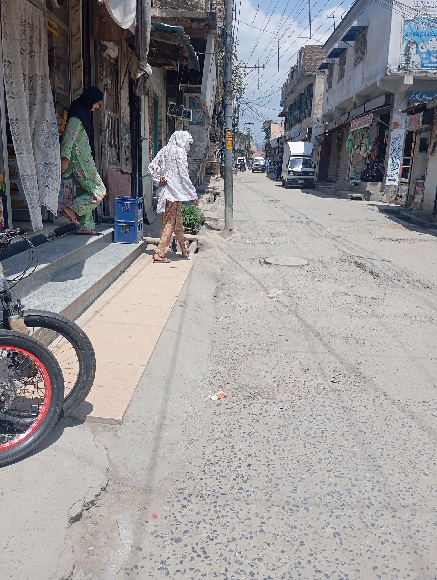 In Mansehra, Pakistan, a street scene unfolds with two individuals on the right sidewalk. One person, wearing a white headscarf and patterned trousers, walks away from the viewer. The second person, dressed in a green patterned outfit and a dark headscarf, stands on a set of steps to the left. The steps lead up to a storefront with white lace curtains. In the foreground, a bicycle with red-rimmed wheels is parked on the lower left. The street itself is paved with asphalt and shows signs of wear, with patches of gravel and some unevenness. Buildings line both sides of the street, featuring awnings and a variety of signage. Power lines crisscross overhead, and a white delivery van is parked further down the street. The sky is partly cloudy.