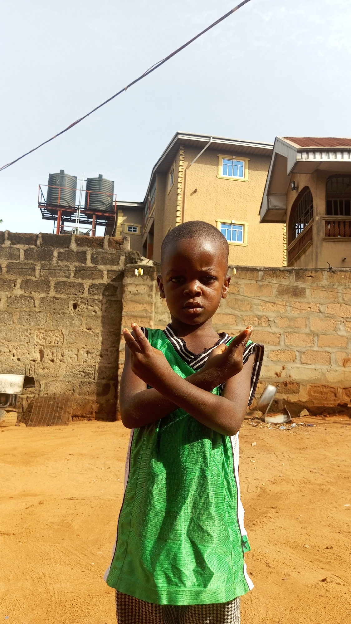 A young boy stands outdoors in Nkpor, Nigeria. He has dark skin and short, cropped hair. He is wearing a green jersey with white and black trim, and plaid pants. He has his arms crossed, with his hands held up and fingers extended in a gesture.
Behind the boy is a brick wall with two black water tanks on a metal frame. Beyond the wall is a tan two-story building with blue-framed windows and a balcony. The ground is a mix of dirt and sand. The sky is pale and overcast.