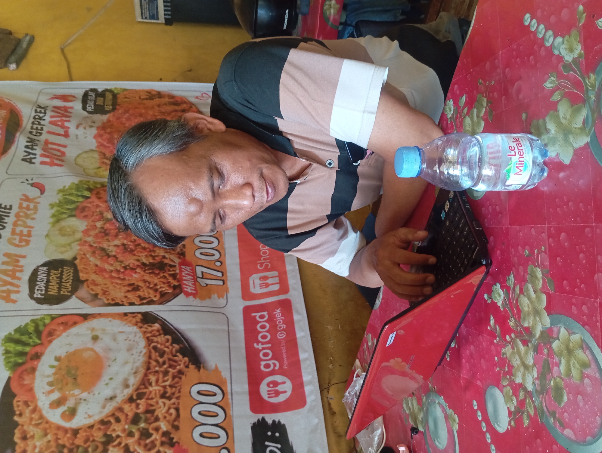 A middle-aged Indonesian man with short, graying hair and a visible lump on his forehead is seated at a red, floral-patterned table, looking down at a red laptop. He is wearing a collared shirt with horizontal stripes of brown, dark green, and white. A bottle of "Le Minerale" bottled water with a blue cap sits to the right of the laptop. Behind the man, a banner displays images of Indonesian food, including "AYAM GEPREK HOT LAVA" and "NASI GORENG". The background appears to be an indoor setting with a yellow wall and part of a black motorcycle helmet visible. The overall environment suggests a small eatery or food stall in Sedati Agung, Indonesia.