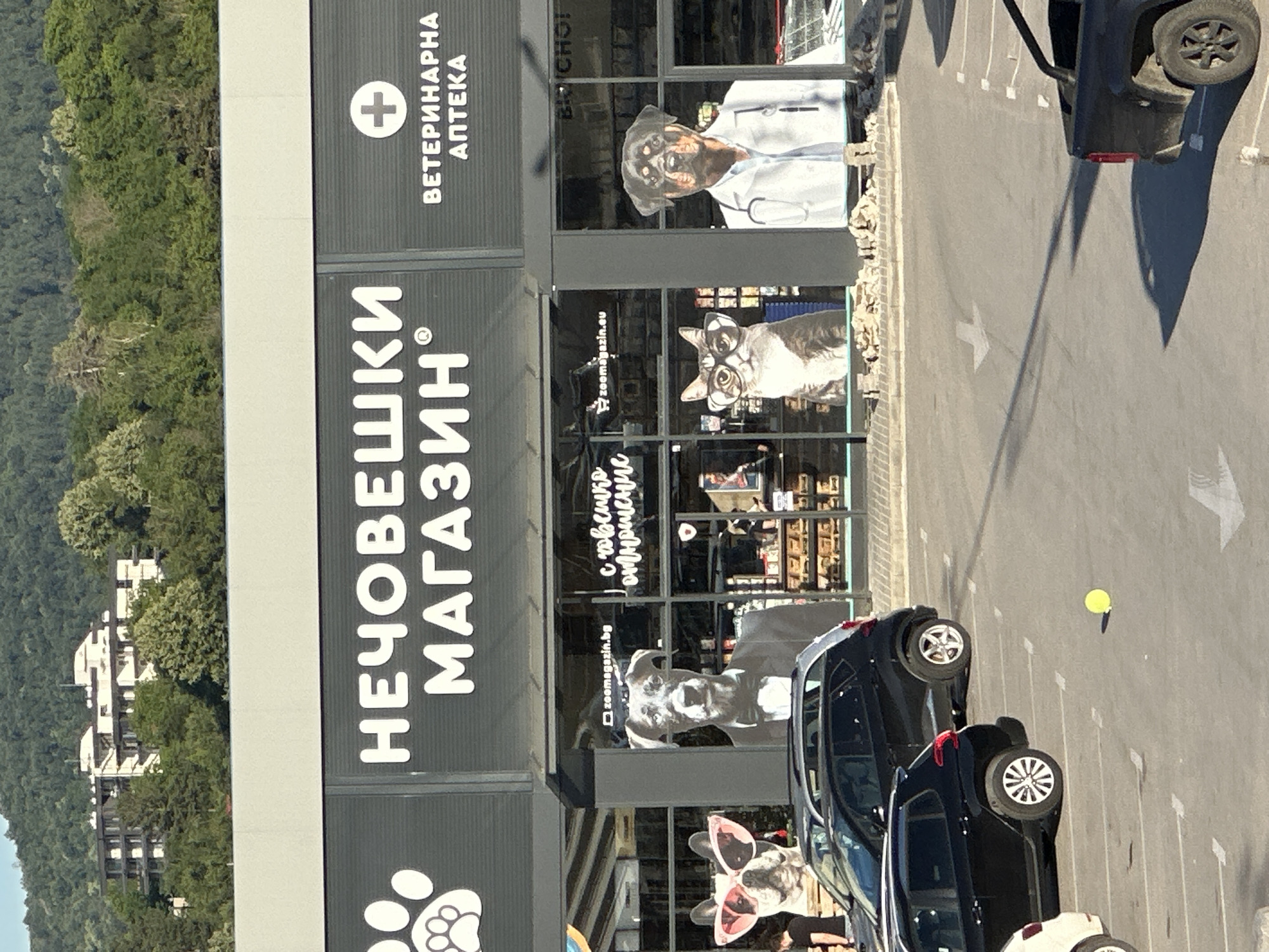 The image shows the exterior of a pet store and veterinary clinic in Veliko Tarnovo, Bulgaria. The store's signage features large white text in Cyrillic, reading "НЕЧОВЕШКИ МАГАЗИН" (Pet Store) and "ВЕТЕРИНАРНА АПТЕКА" (Veterinary Pharmacy). The facade is predominantly black and gray, with large windows displaying graphics of animals. Specifically, there are large cutouts of a dog wearing sunglasses and a tuxedo, a cat wearing glasses, and a Rottweiler dressed as a veterinarian. The store appears to be a modern commercial building situated in a suburban or semi-rural area, with a backdrop of a forested hill and some residential buildings. In the foreground, a parking lot is visible with several parked cars, including black SUVs and a white car. A yellow ball is on the ground in the parking lot. The lighting suggests it is daytime with clear weather.