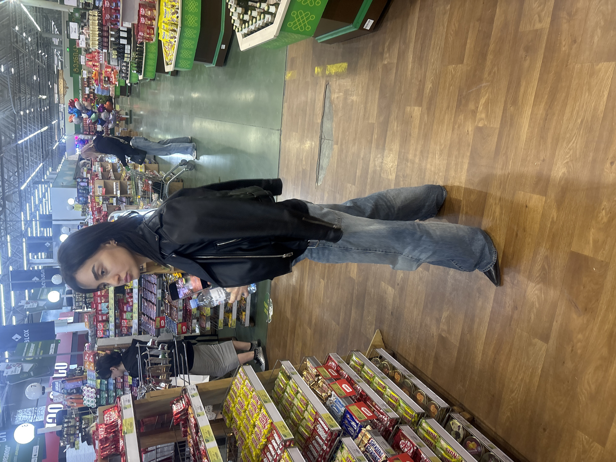 The image shows a person standing in a supermarket aisle. The person is wearing a black leather jacket and blue jeans. They are looking at the shelves. The shelves are stocked with various candy bars, snacks, and canned goods. Other shoppers are visible in the background. The location is likely in a city, and the signage in the image suggests it could be Tbilisi, Georgia.