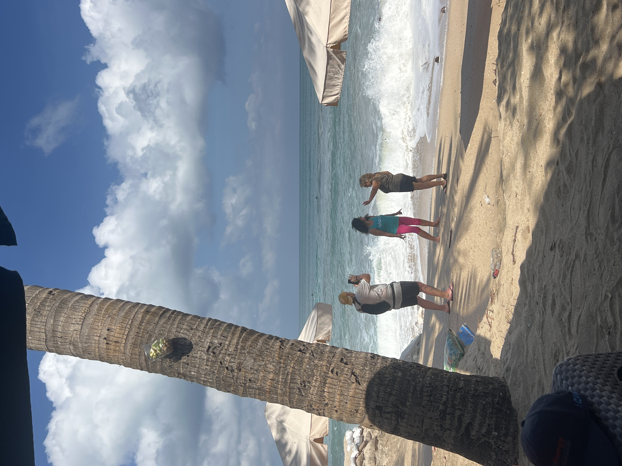 In Kuta, Indonesia, a palm tree trunk dominates the left foreground of a beach scene. Attached to the trunk is a clear plastic bottle filled with light-colored objects, possibly food. In the middle ground, three people are on the sandy beach. A person wearing a hat and backpack faces the ocean, holding a mobile phone as if taking a photo. To their right, two women stand facing each other near the water's edge, with waves crashing on the shore. One woman wears a teal tank top and pink leggings, while the other wears a patterned top and dark shorts. Two large, cream-colored beach umbrellas are visible behind the people, and in the distance, the ocean stretches to the horizon under a blue sky with scattered clouds. The sand is strewn with debris, including a blue bag and a clear plastic bottle. In the bottom left corner, a dark baseball cap rests on a woven chair, casting a shadow.