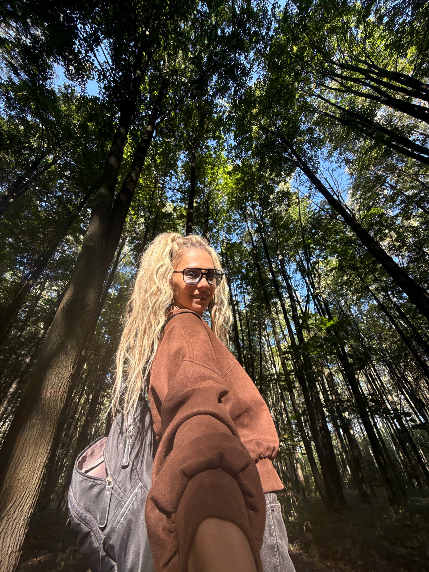 A low-angle, wide shot captures a young Caucasian woman with blonde, curly hair, wearing large dark sunglasses, a brown sweatshirt, and jeans, standing in a dense forest with tall trees. She has a black backpack slung over her left shoulder and is holding her right arm up, as if taking a selfie. The sunlight filters through the leaves of the trees, creating dappled light and shadows on the forest floor and the woman's body. The trees are predominantly deciduous, with thick trunks and abundant green foliage. The sky is visible between the branches, appearing a bright blue. The scene is set in the city of Rusanovo, Russia.