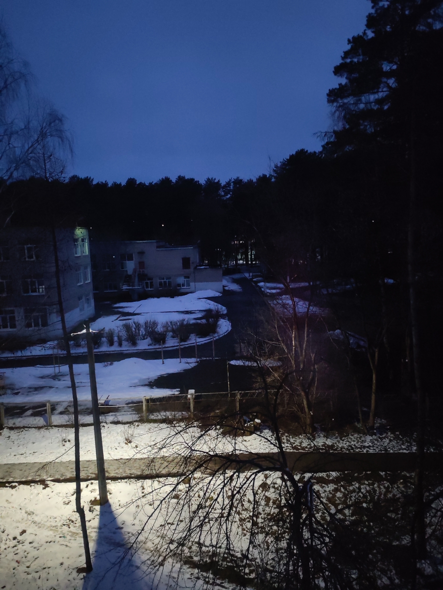 The image shows a winter scene in Chaikovsky, Russia at dusk or night. There are snow-covered grounds, bare trees, and a two-story building with lit windows in the background. The buildings appear to be part of a residential or institutional complex. There is a road or pathway leading to the buildings. A dark forest line is visible in the distance. The foreground features a sidewalk and leafless branches, suggesting a low vantage point, possibly from a higher story of a building. There are no people visible in the shot. The overall atmosphere is dim and still.