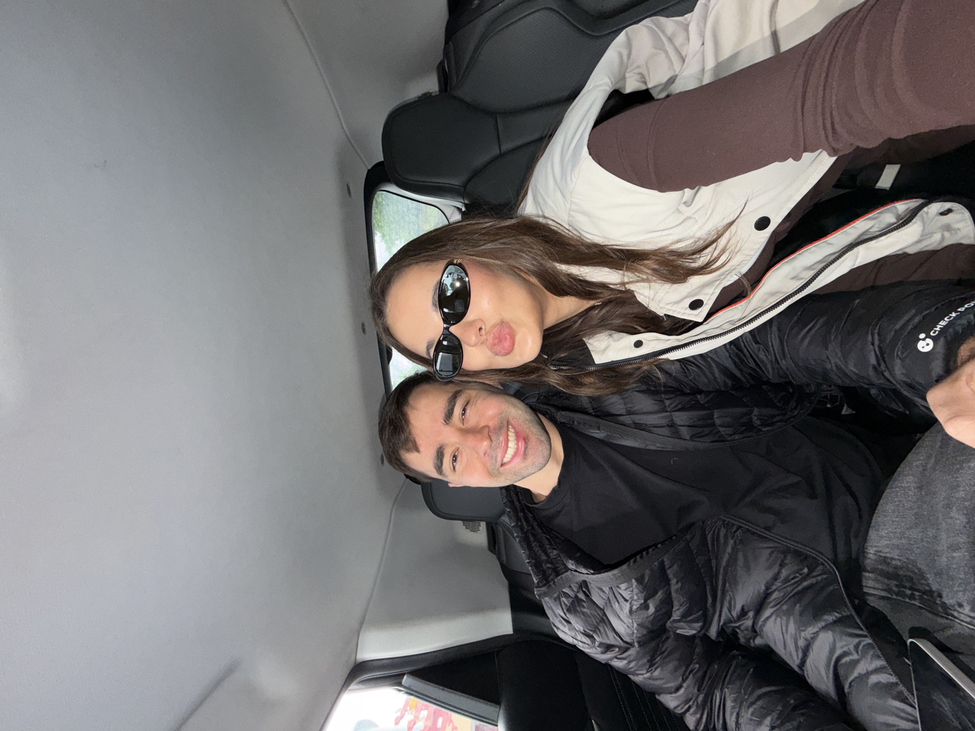 Inside a vehicle, a man and a woman are captured from an elevated angle. The woman is wearing sunglasses and puckering her lips, while the man is smiling. The woman is wearing a white jacket over a darker garment. The man is wearing a black quilted jacket over a black shirt. The background reveals the interior of the vehicle, including seating and the car's ceiling. The setting appears to be Yongding District, China, as indicated by the presence of possible Chinese characters outside the window.