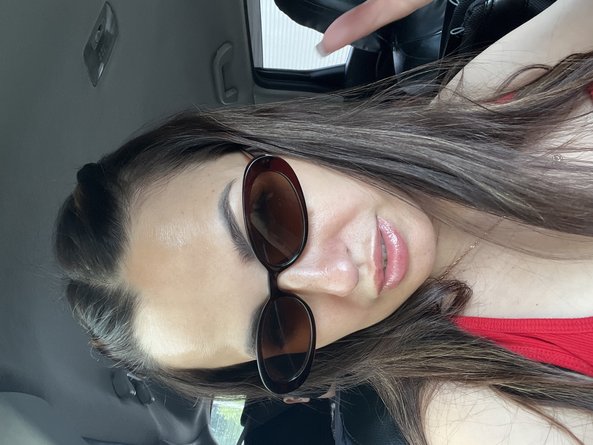 A woman is captured in a close-up, front-facing selfie taken from inside a vehicle. She is wearing dark-rimmed, oval sunglasses that obscure her eyes. Her hair, dark brown with subtle lighter streaks, is styled in a half-up, half-down fashion, with two small buns at the crown of her head. She has on a red top, and a delicate gold necklace is visible around her neck. Her lips are glossy and slightly parted, and she is making a peace sign with her right hand, which is positioned in the upper right portion of the frame. The background suggests the interior of a car, with a dark headliner, a door handle, and tinted windows visible. The lighting indicates it is daytime, and the overall atmosphere is casual. The specific location within the city of Unknown, Unknown cannot be determined from the image.
