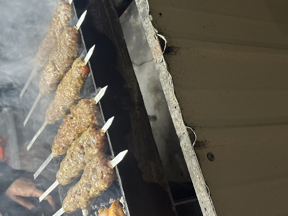 Six skewers of ground meat, possibly kebabs, are actively grilling over a charcoal fire, emitting visible white smoke. The meat on the skewers appears to be minced or ground, with some browning evident on the surfaces. A seventh, smaller, irregular food item, lighter in color, is visible on the far left of the grill. The skewers rest on a dark, heavy-gauge metal grill structure, likely a mangal. A person's hand, partially visible in the upper left, wearing a dark sleeve, is present near the cooking area. Below the main grill structure, a section of a light-colored, possibly corrugated metal or concrete barrier is visible, showing signs of wear and a single screw attachment. The scene is located in the city of Pap, Uzbekistan.