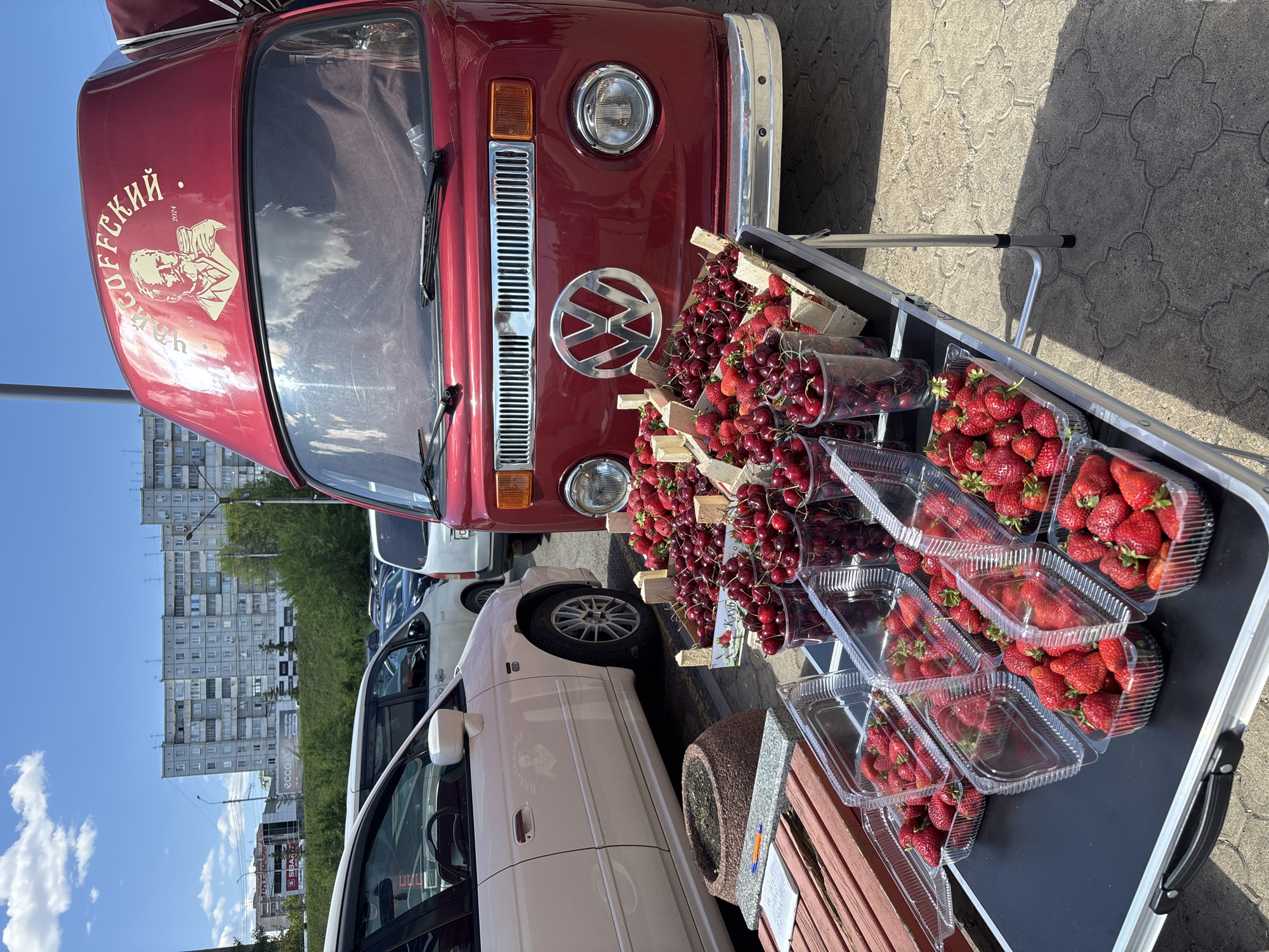 The image shows a street-side fruit stand in Krasnoyarsk, Russia. The foreground features a table laden with containers and baskets of fresh strawberries and cherries. A red Volkswagen van with a white sticker featuring a man and the inscription "OFFSKI 2024" stands directly behind the table, partially obscured by the fruit display. A white sedan is parked to the left of the van. In the background, a multistory apartment building rises into a bright blue sky with scattered clouds. Other vehicles are parked along the street. The pavement has a patterned texture.