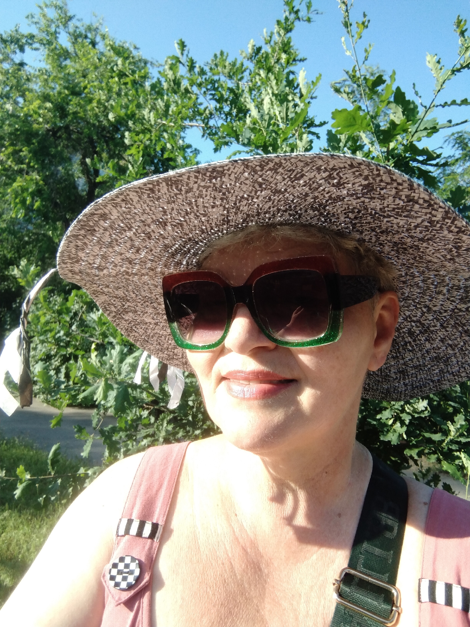A woman is pictured outdoors, wearing a wide-brimmed straw hat, oversized sunglasses with green and brown rims, and a pinkish-red garment with a dark green strap. Her left shoulder and upper chest are visible, as are parts of her face including her mouth and chin, which are illuminated by sunlight. She has short, blonde hair. The background consists of lush green trees and a clear blue sky. The image appears to be taken in a park or garden setting. There is no visible indication of the specific location within Horishni Plavni, Ukraine, other than the implicit assumption it is taken there.