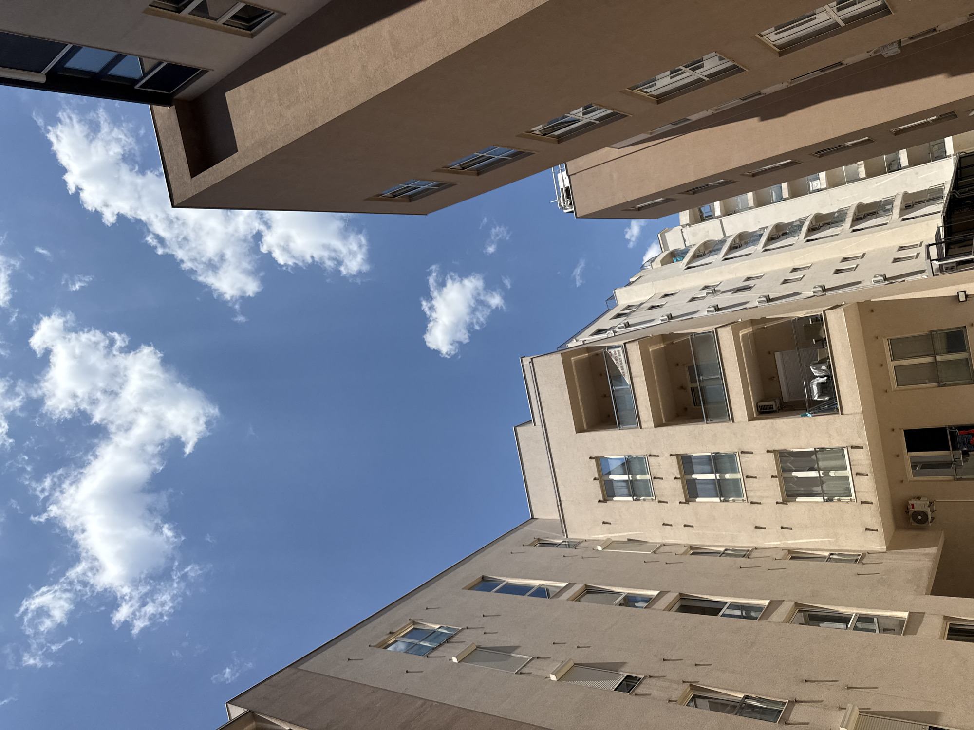 This is a low-angle, full shot of apartment buildings in Budva, Montenegro, against a bright blue sky with scattered white clouds. The buildings are modern, with stucco exteriors in neutral tones of beige and brown.
The building on the left is lighter in color and has multiple windows with white frames, some with closed roller shutters. There are small, vertical protruding elements spaced evenly across its facade. Air conditioning units are visible on several floors.
The building in the center is also beige, with a more ornate design featuring balconies with glass railings and curved edges. More windows and air conditioning units are visible.
The building on the right is taller and a darker brown color. It has a simpler, more angular design with rows of tall, narrow windows. The building casts a shadow on the beige building to its left.
The sky is a clear, vibrant blue, with fluffy white cumulus clouds that are illuminated by the sun. The perspective is looking upwards, emphasizing the height of the buildings and the expanse of the sky. The overall impression is one of urban residential architecture under a clear, sunny sky. There are no people visible in the image.