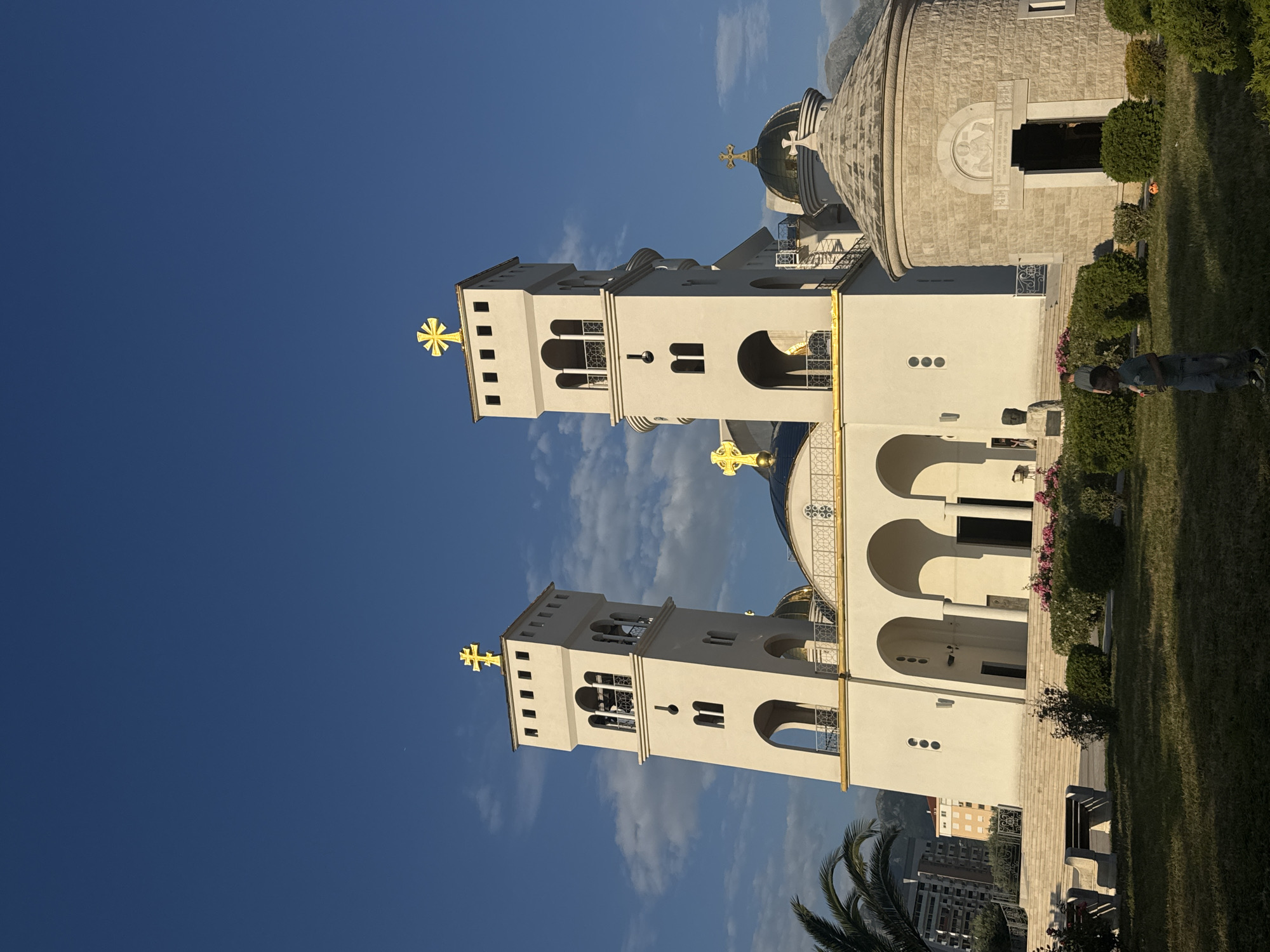 The image captures the Church of St. John Vladimir in Bar, Montenegro. The church features two prominent, white bell towers topped with golden crosses. A rounded, stone building is adjacent to the towers, also bearing a golden cross atop its dome. The sky above is a clear blue, with some scattered clouds visible behind the towers. The foreground includes manicured green lawn and some shrubbery. To the right, a person stands on the grass. In the distance, building structures are visible.
