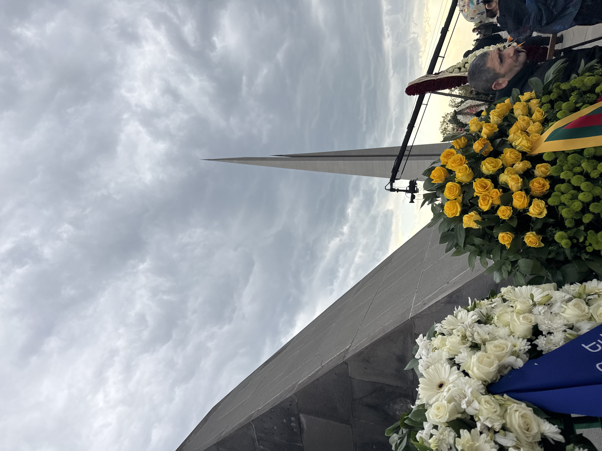 The image captures a solemn outdoor scene at what appears to be a memorial site, likely the Tsitsernakaberd Armenian Genocide Memorial in Yerevan, Armenia, given the context and distinctive architecture. The central feature is a tall, slender, dark grey pointed stele dominating the left side of the frame, reaching towards a heavily overcast sky filled with grey clouds.
In the foreground on the right, two large floral wreaths are prominently displayed. The upper wreath is composed primarily of vibrant yellow roses and dense green foliage, possibly chrysanthemums. A ribbon showcasing the horizontal yellow, green, and red stripes of the Lithuanian flag is visibly integrated into this wreath. Partially visible below this is another wreath made of white flowers, including roses and gerbera daisies, adorned with a blue ribbon on which the partial text "TU" or "TUL" can be seen.
Several people are gathered behind the wreaths. A man with short grey hair and a beard, wearing dark clothing, is visible in profile, looking upwards or into the distance with a contemplative expression, suggesting a moment of remembrance or reflection. A microphone on a stand is also present, indicating a formal ceremony or a public address. The cloudy sky contributes to a somber and subdued atmosphere, aligning with a commemoration or memorial event. The overall mood conveyed is one of respect, solemnity, and remembrance.