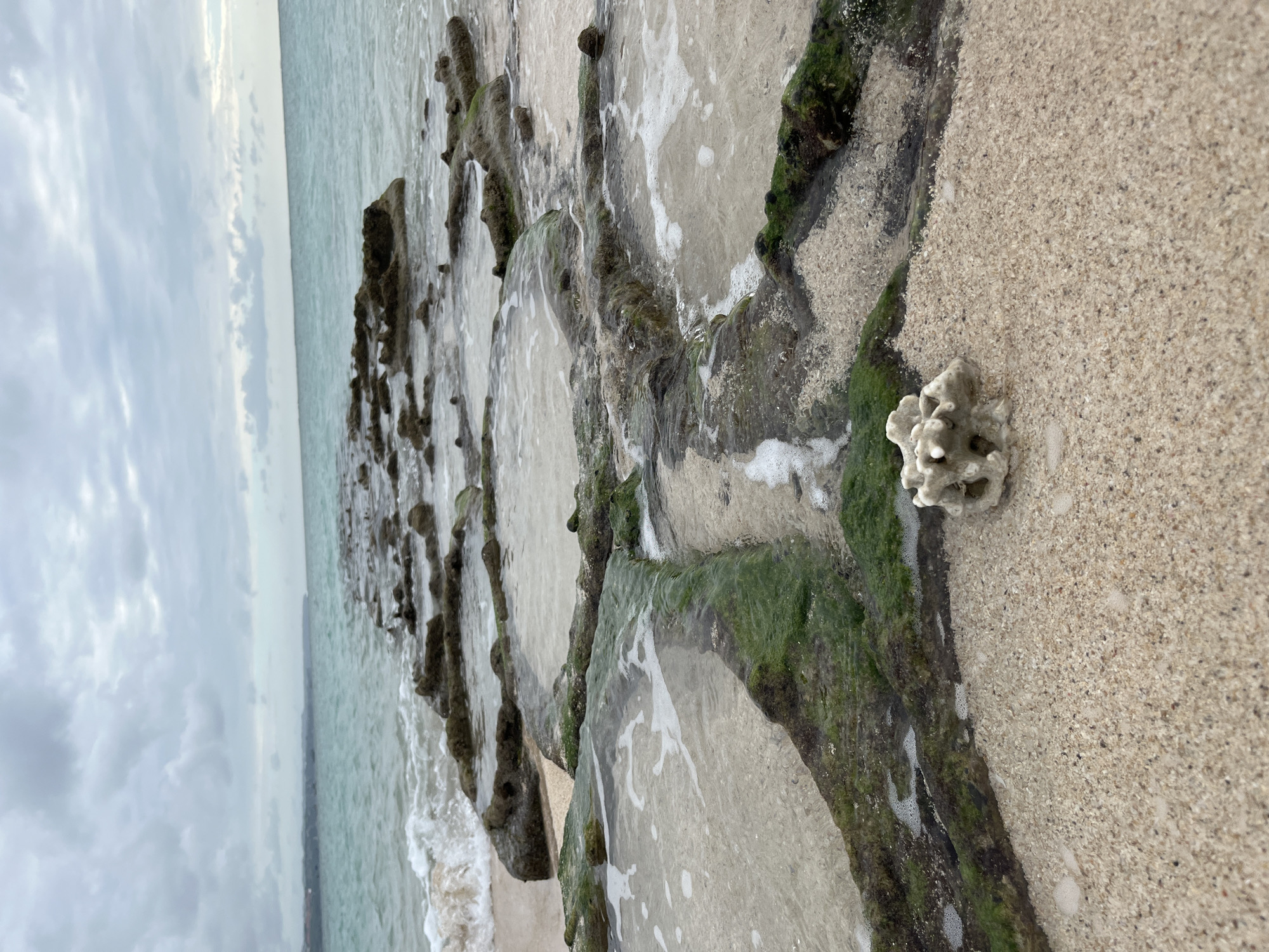 This is a close-up, eye-level, vertical shot of a rocky shoreline on an overcast day. In the foreground, a small piece of bleached coral sits on tan sand. Extending into the ocean is a line of dark, rough-textured rocks that appear to have been carved or naturally formed into a linear pattern, some with shallow pools of water reflecting the sky. The water is a murky turquoise, with small waves lapping at the shore. The background shows a distant, hazy coastline under a cloudy sky. The scene is likely a natural formation or a man-made structure of some kind, possibly for erosion control or fishing. The location is Kuta, Indonesia.