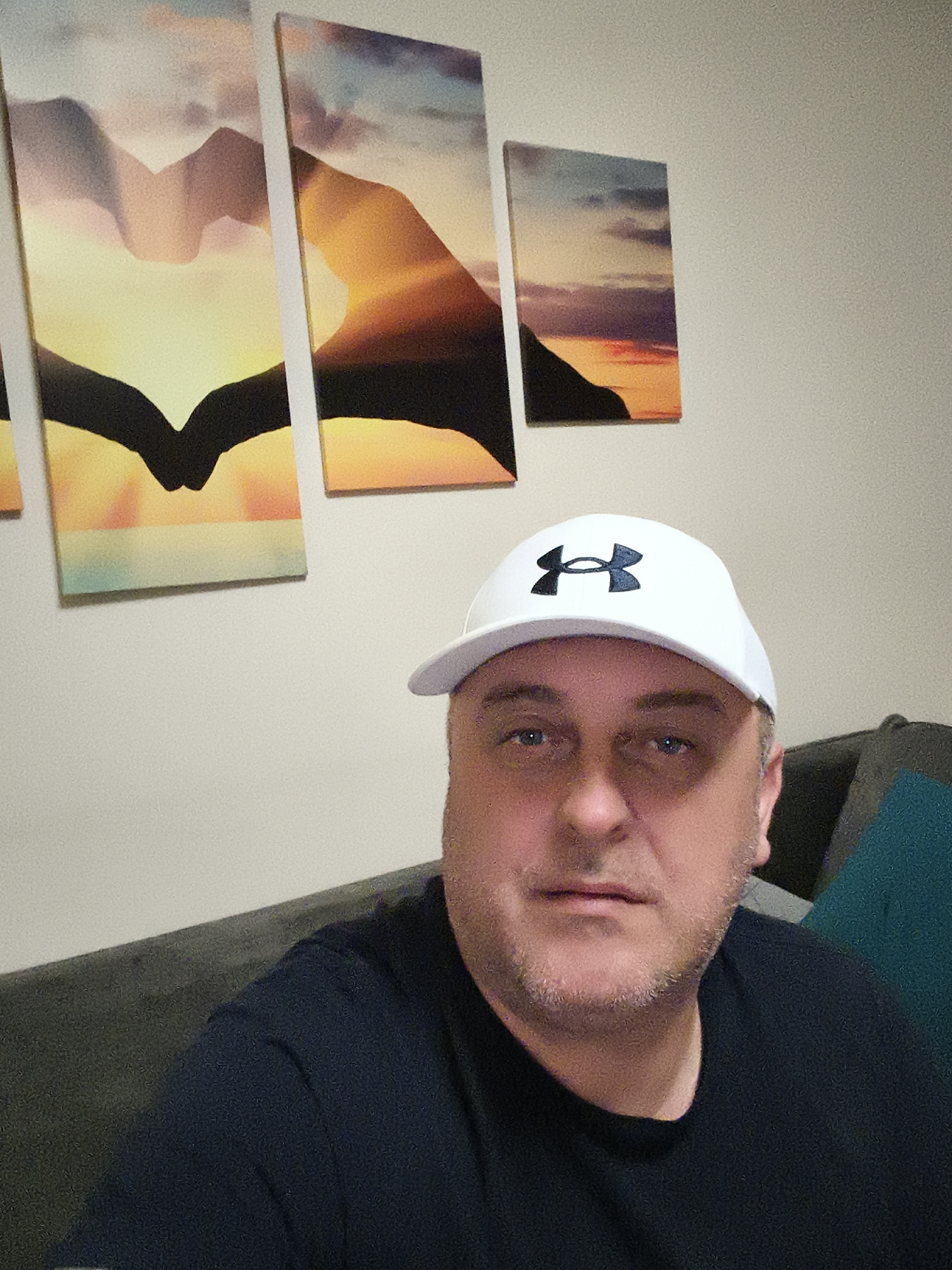 A man, appearing to be middle-aged with fair skin, blue eyes, and light facial stubble, is depicted in a casual indoor portrait. He is wearing a white baseball cap with the black, stylized "H" logo of Under Armour visible on the front, along with a dark, likely black or navy, short-sleeved t-shirt. The man looks directly at the camera with a neutral expression.
He is seated, with a portion of a grey or dark-colored sofa visible on the left and a teal or turquoise cushion visible on the right. Behind him, mounted on a plain, light-colored wall, is a collection of four canvas prints. The main three prints, arranged in a triptych, depict a stylized sunset or sunrise over mountain-like silhouettes, with vibrant yellows, oranges, and purples blending into blues and greys in the sky. The dark shapes at the bottom of these three panels converge to form a distinct heart-like or bird-in-flight silhouette. A fourth, smaller panel to the right continues the sunset/mountain theme.
The overall setting suggests a domestic environment, such as a living room, located in Huntingdonshire, United Kingdom. The image has a slightly grainy texture. No specific activity beyond posing for the photograph is evident, and the time of day is not discernible from the indoor lighting.