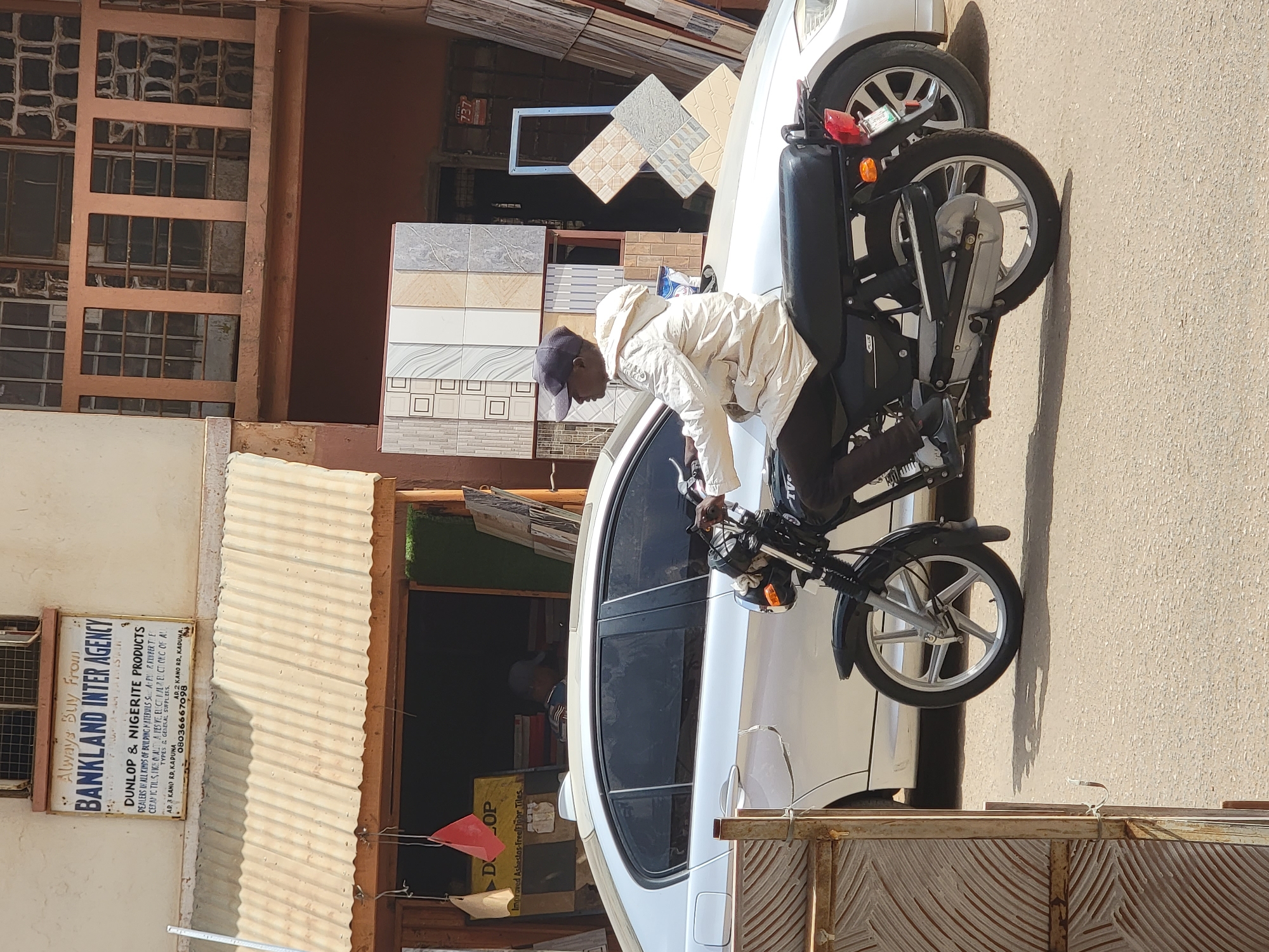 A person on a black scooter is parked on the side of a white car in Maiburiji, Nigeria. The scooter is parked in front of a building supply store, "BANKLAND INTER AGENCY", a dealer of "DUNLOP & NIGERITE PRODUCTS", with tile displays. The scooter driver has a light jacket and a dark baseball cap. The background shows buildings and the street.