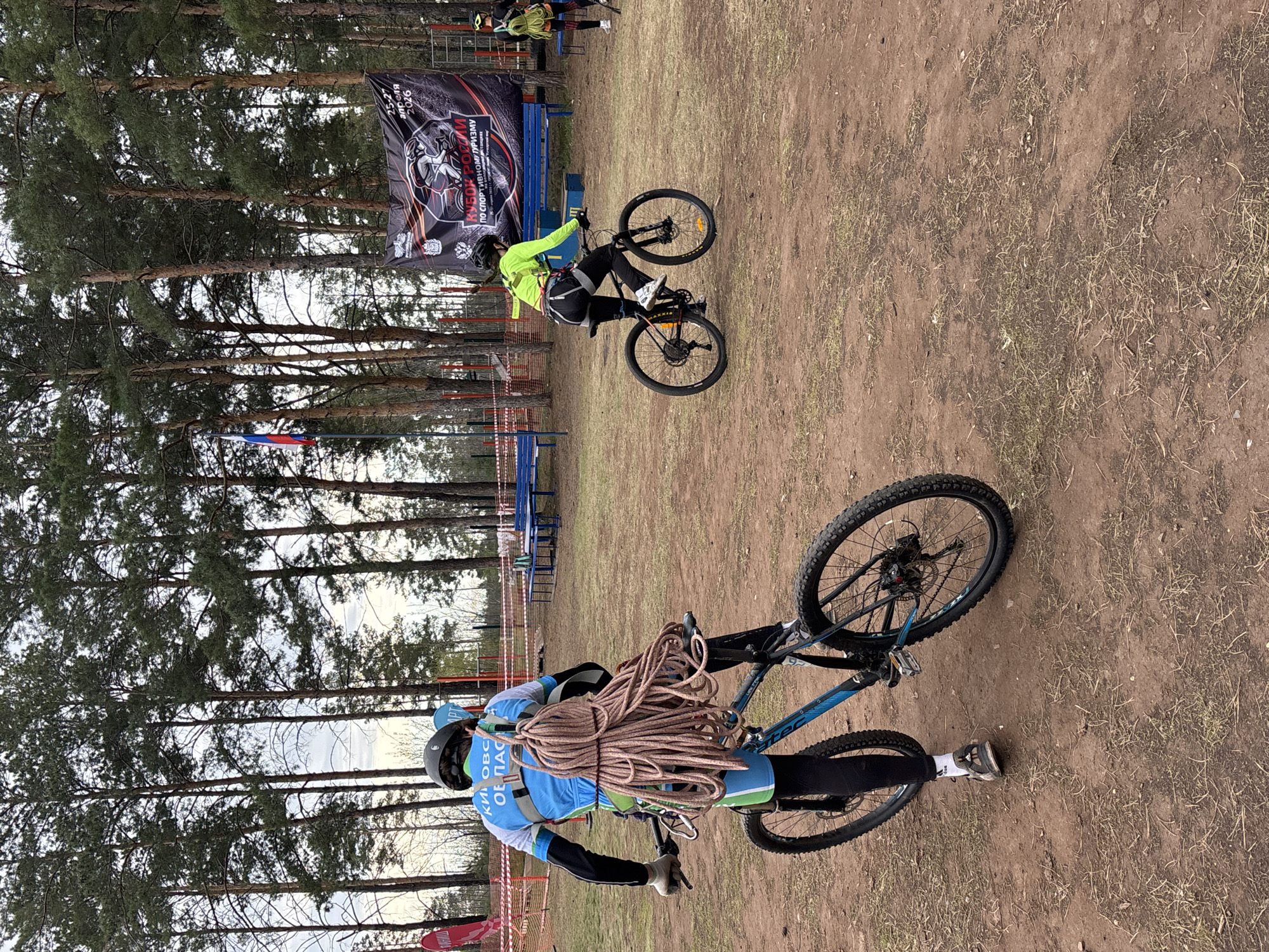 The image depicts an outdoor scene in what appears to be a park or wooded area, possibly in the city of Orenburg, Russia, given the Russian text and flag. The ground is dirt with sparse vegetation. Tall trees are visible in the background.
Two individuals on bicycles are present. One is wearing a blue and green outfit with a helmet and carries a large rope on their back. The other is wearing a bright yellow jacket and black pants, riding a bike.
There's a banner in the background with Russian text. Blue benches are also visible in the background. Red and white striped tape marks the perimeter of an area. The overall setting suggests some kind of outdoor event or competition involving biking and possibly climbing or other outdoor skills.