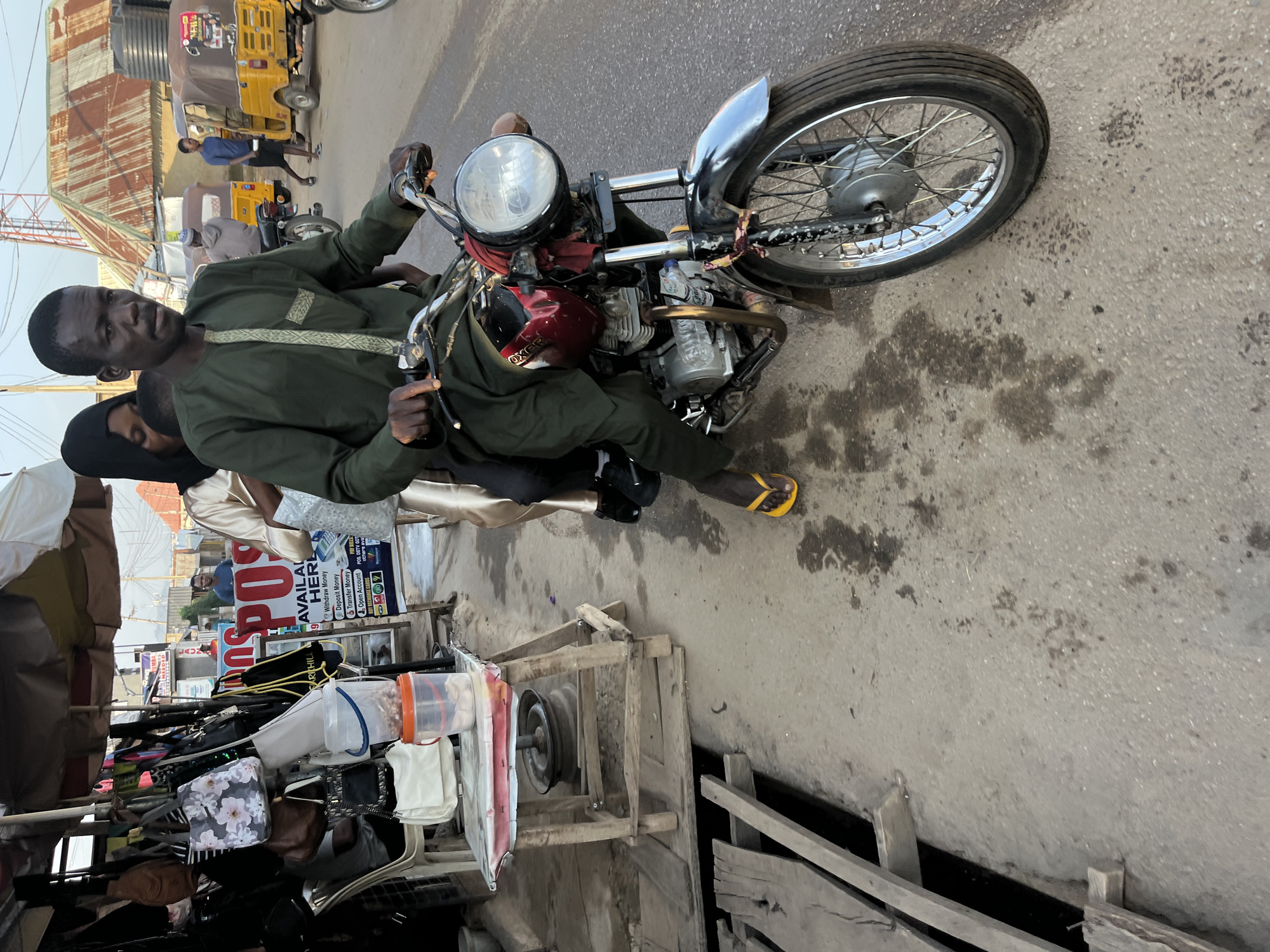 In Abuja, Nigeria, a man and a woman ride on a motorcycle down a street, with the man driving and the woman behind him. They are dressed in traditional clothing. In the background, there are market stalls and other vehicles, including a yellow tricycle. The street is paved with asphalt and there are some puddles of water on the ground. The overall impression is that of a busy street scene in a Nigerian city.