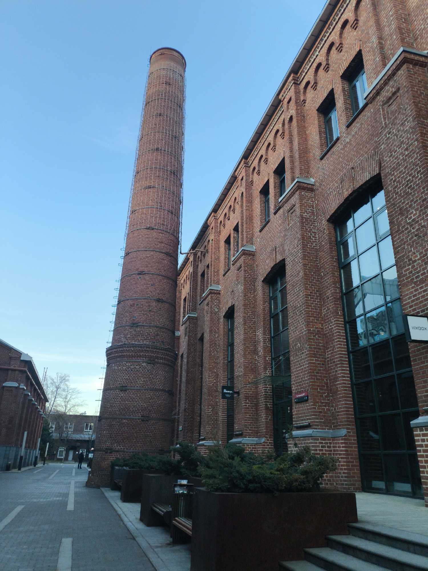 This image presents a striking view of an urban landscape in Moscow, Russia, characterized by industrial heritage architecture. The central subject is a towering red brick chimney, reinforced with metal bands and featuring a ladder-like structure along its side, standing prominently against a clear, bright blue sky.
Adjacent to the chimney, a long, multi-story brick building dominates the right side of the frame. This building showcases intricate red brickwork with decorative arched details above its numerous large windows and along its roofline. The windows themselves are expansive, reflecting the blue sky.
A narrow paved street or pedestrian path runs between the chimney and another, less prominent brick building on the left. In the distance along this path, a solitary person dressed in dark clothing can be observed walking away, contributing to a sense of quietude. Modern, dark metal planters filled with evergreen shrubs line the path near the main building, alongside benches made from similar materials, indicating a recently revitalized area. Concrete steps lead up to entrances of the main building.
The time of day appears to be either a crisp morning or late afternoon, given the clear blue sky and the quality of light. Bare trees in the background suggest cooler weather or winter.
Visible text includes "KIKBOX" on a white sign affixed to the main brick building on the right. Fainter signs further down the building read "LA MERCE..." and possibly "HOORAYS". A small red sign with the number "22" and an arrow pointing left is also visible. The overall scene evokes a blend of historical preservation and contemporary urban renewal, presenting a calm and composed atmosphere.