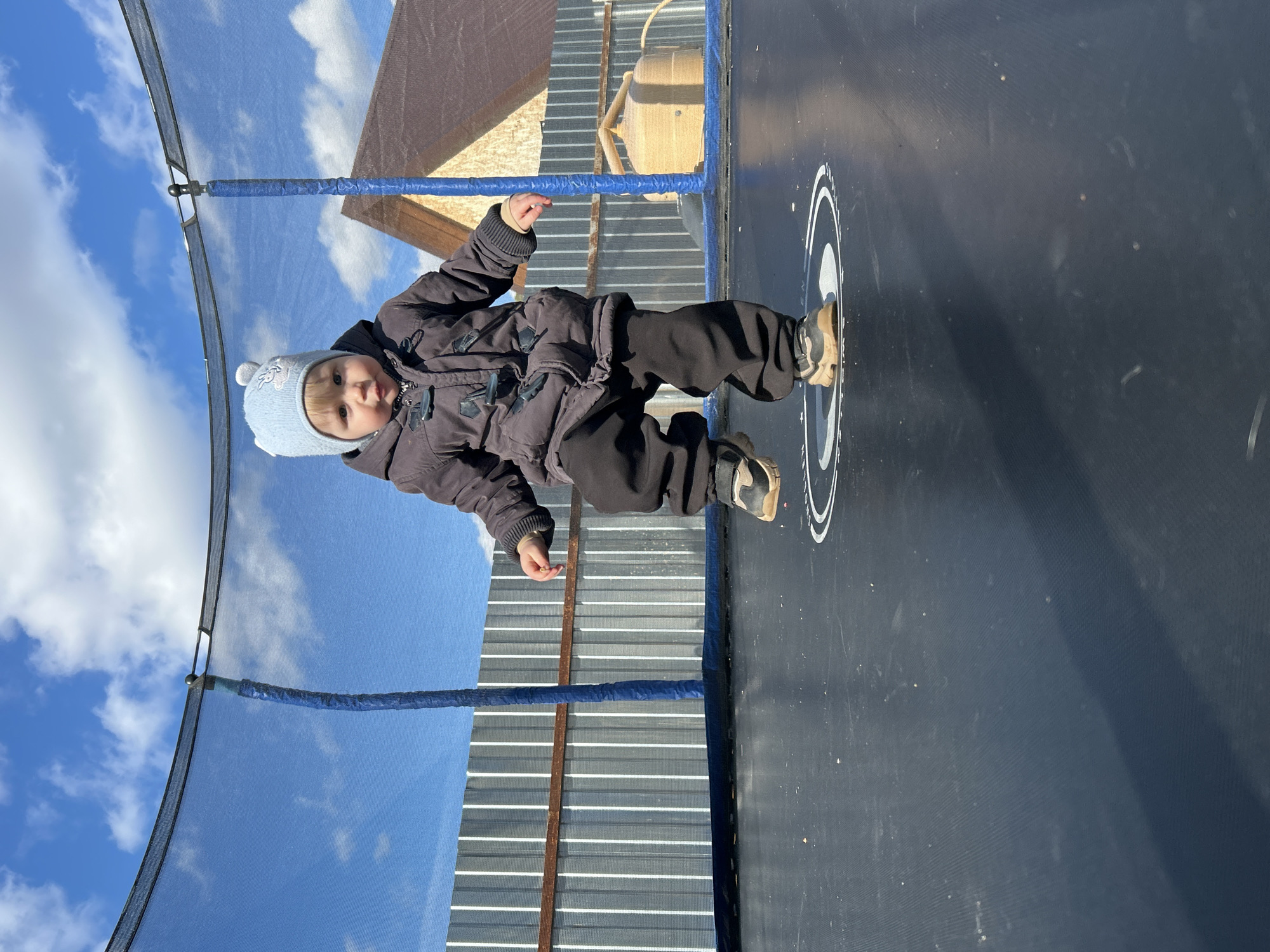 A young child is on a trampoline in an outdoor setting. The child is wearing a dark-colored winter coat, dark pants, and a light blue knit hat. The trampoline has a safety net enclosure around it. The background includes a cloudy blue sky and a corrugated metal fence. Part of a building with a light brown roof is also visible. The location is Chaikovsky, Russia. The child is seemingly in mid-air, likely jumping on the trampoline.