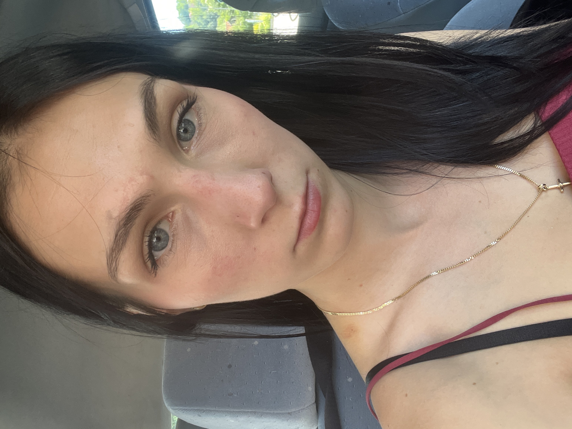 A young woman with long, dark hair is captured in a selfie taken inside a vehicle. She is wearing a red and black strap top and a gold chain necklace. Her facial expression is neutral, with her gaze directed towards the camera. The background shows the interior of a car, with a headrest visible on the left and a blurred view of trees and buildings through the window on the right. The lighting suggests it is daytime.