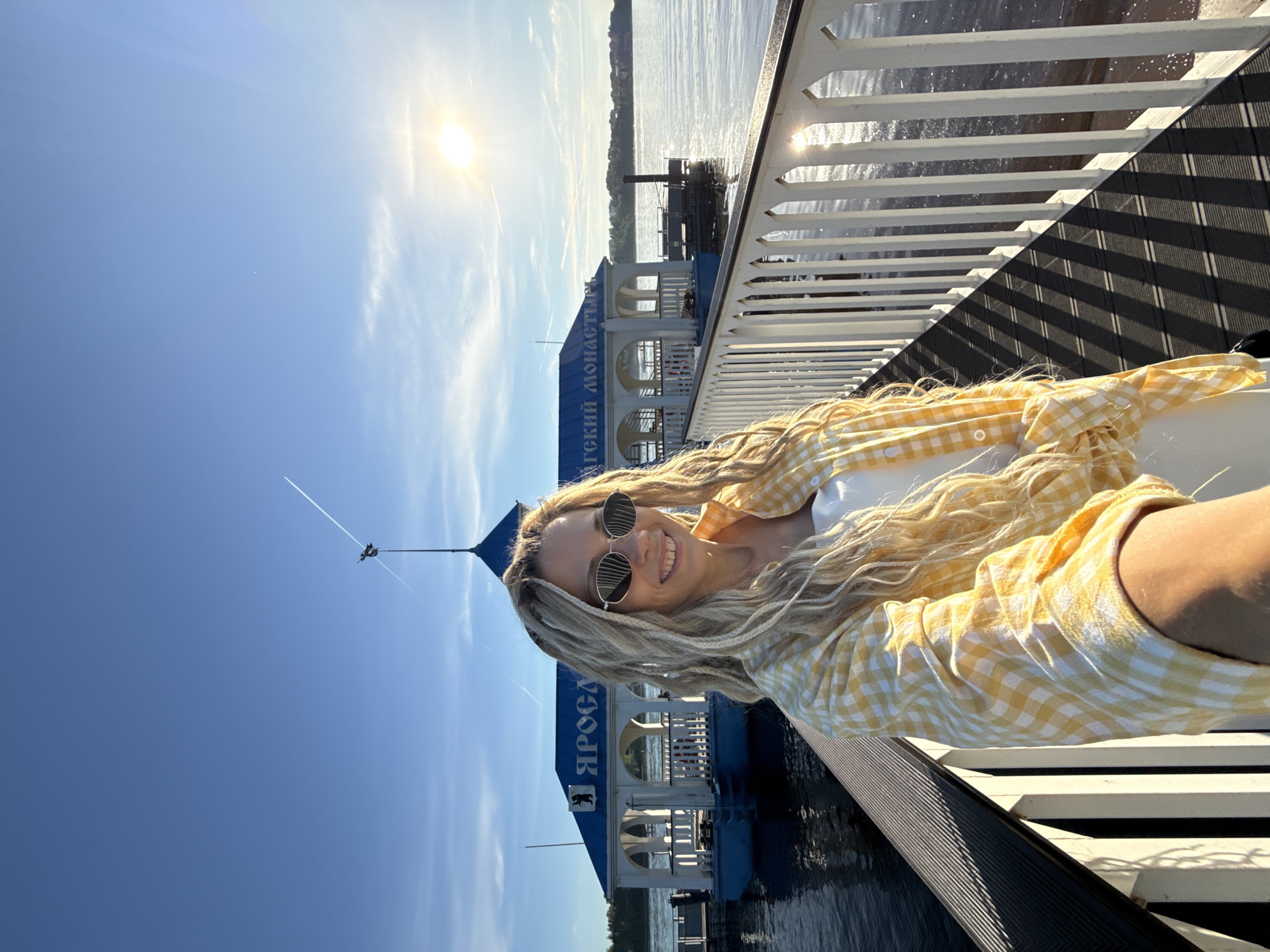 A sunny outdoor scene depicts an adult female capturing a selfie on a pier adjacent to a body of water in the Dumino area, Yaroslavl Oblast, Russia. The woman, positioned centrally, has long, blonde dreadlocks or braids and wears dark, hexagonal sunglasses, a white top, and an open yellow and white checkered long-sleeved shirt. She smiles directly at the camera, holding it in a self-portrait orientation with her right arm extended into the lower right foreground.
The pier features a dark, grooved deck and a white decorative railing on the right, casting long, parallel shadows across the deck due to the low angle of the sun. The body of water, likely the Volga River, is visible on both sides of the pier, reflecting bright sunlight intensely on the right side.
In the midground, a white pavilion-style building with a distinctive blue, peaked roof is docked to the pier. The building has multiple arched openings and features partially visible Russian text on its façade: "ЯРОСЛА" (Yarosla-) on the left and "-ГСКИЙ МОНАСТЫРЬ" (-gskiy Monastyr) on the right, indicating a connection to the Yaroslavl Monastery or River Station in Yaroslavl. A dark bear emblem, consistent with the coat of arms of Yaroslavl, is visible above the "ЯРОСЛА" text. A flagpole with a weathervane in the shape of a creature or bird adorns the peak of the blue roof. Further down the river to the right of this structure, another smaller, dark pier or docking platform is visible.
The background reveals a clear, bright blue sky with sparse, wispy clouds near the horizon. The sun is intensely bright in the upper right quadrant, creating significant glare. Two distinct white contrails from aircraft are visible in the sky; one diagonal streak extends from the upper left to the weathervane, and another faint one appears in the upper right. A distant, tree-lined shoreline is visible across the water. The overall lighting suggests either early morning or late afternoon.