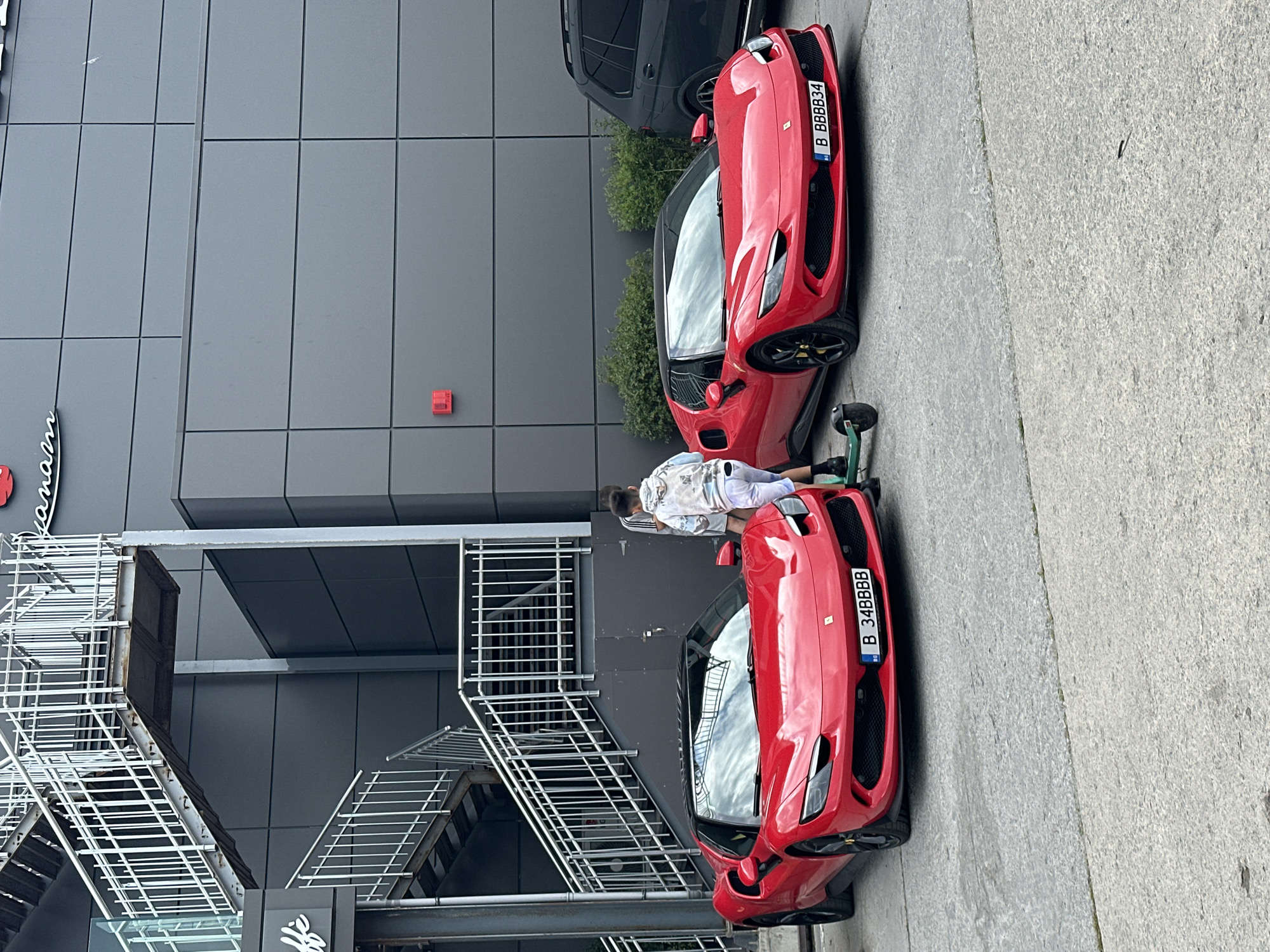 The scene depicts two red Ferrari sports cars parked on a paved surface next to a modern building. A person, dressed in a white-patterned shirt and white pants, is standing between the two cars. A green scooter is also visible near the first car. The building has a gray facade with grid-like paneling and a metal fire escape staircase on the side closest to the camera. The license plates on the cars read "B 34BBBB" and "B BBBBB34," indicating they are registered in Bulgaria. The presence of a "Mann and" sign, although partially cut off, combined with the overall setting, suggests this photo was taken in the city of Varna, Bulgaria, likely near the Mann and Hummel factory or offices there.