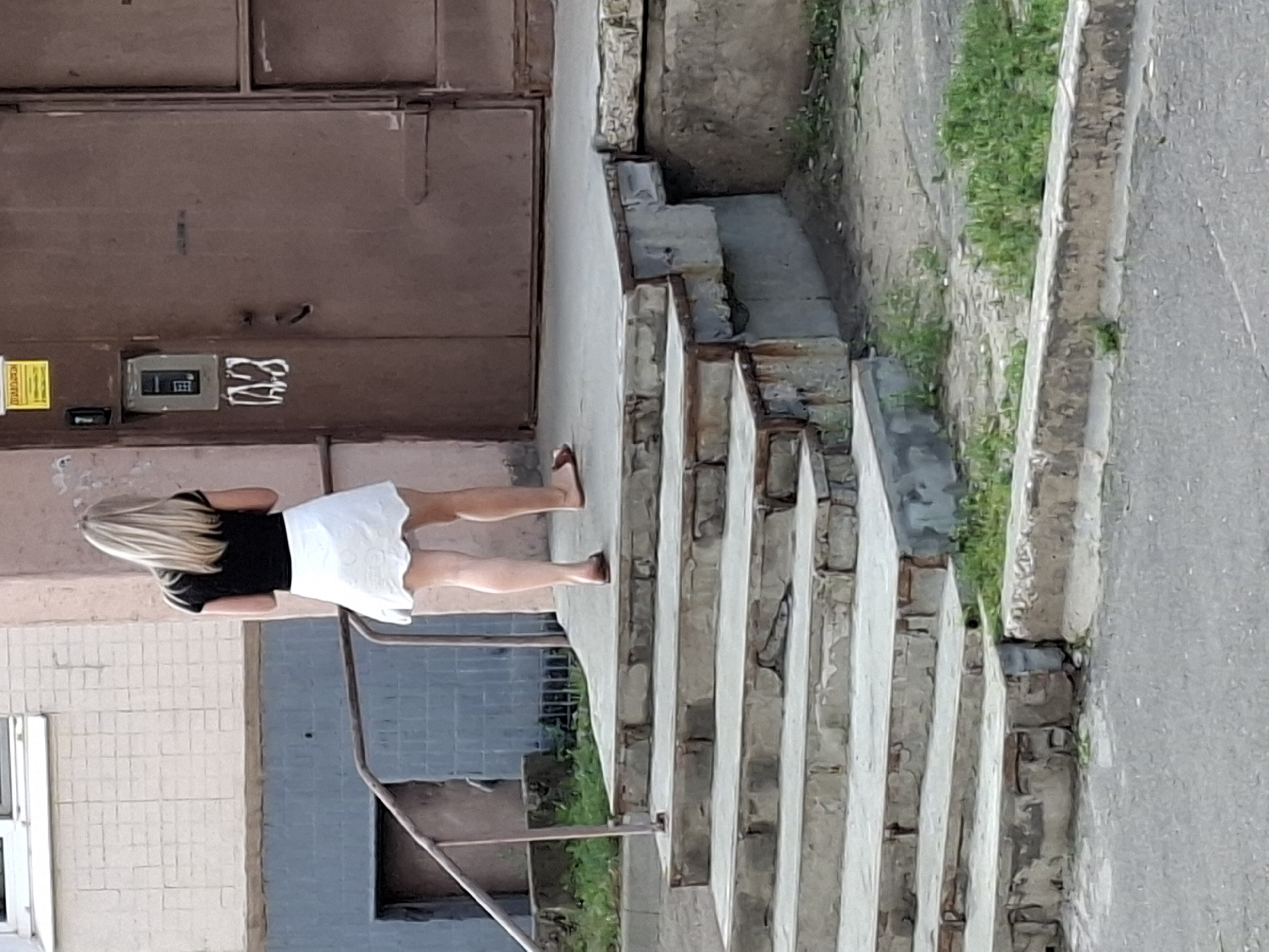 A full-shot image shows a Caucasian woman with blonde hair and a black shirt and white skirt walking up a set of outdoor concrete stairs towards a brown door. The woman is facing away from the camera. The stairs are worn and appear to be made of concrete blocks with metal trim. A metal handrail is visible on the left side of the stairs. The building facade behind the stairs is made of light-colored brick and has a blue tiled lower section. The door is a large, brown metal door with a small window or intercom panel to the left of the door. There is some graffiti visible on the wall next to the door. Green grass and weeds are growing around the base of the stairs and the building. The ground in front of the stairs is asphalt. The sky is not visible in the frame.