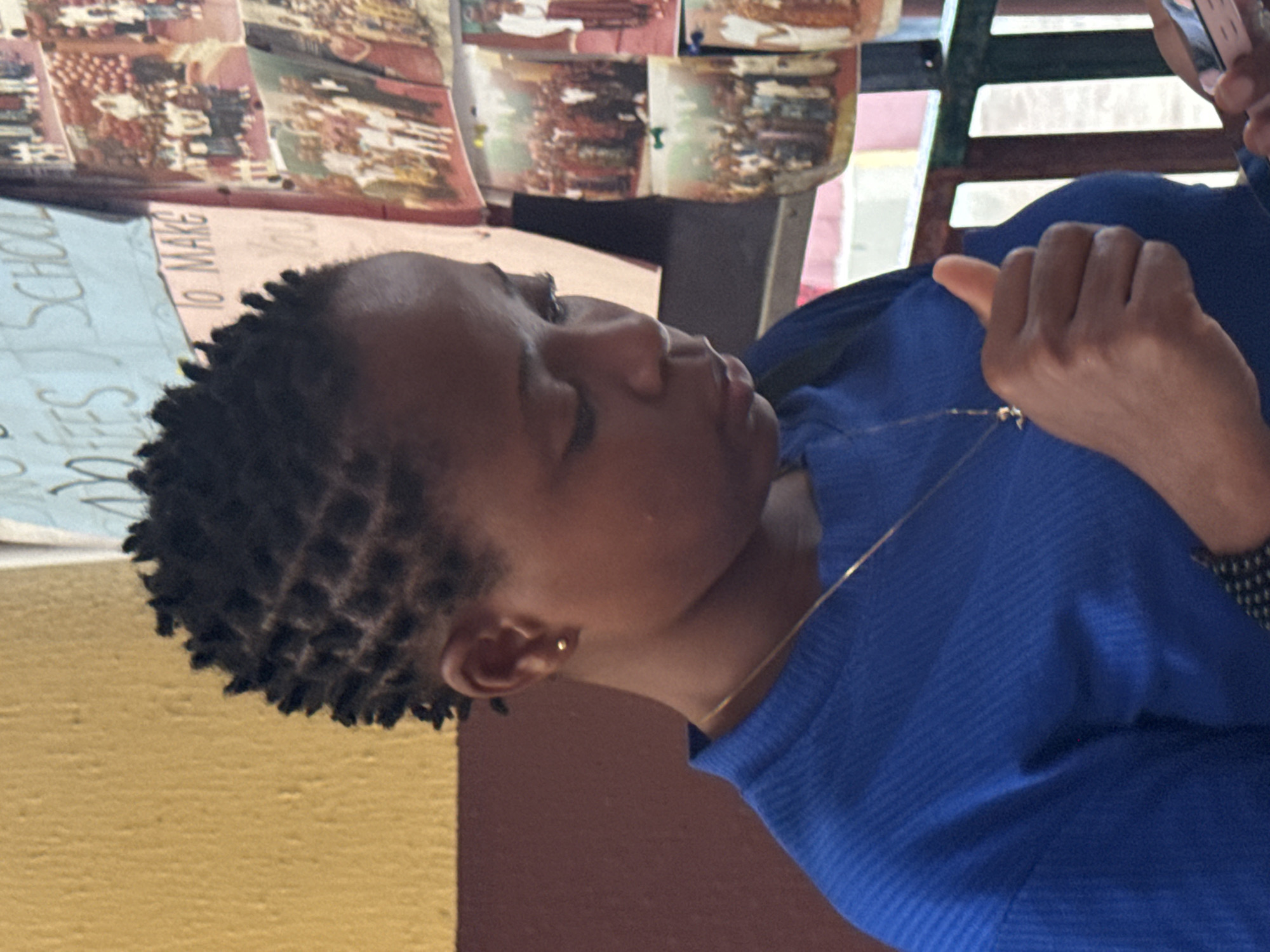 A close-up, profile image captures an individual, likely female, in Ado, Nigeria. The person is positioned on the left, facing right and looking slightly downwards. They have short, dark hair styled in distinct, square-patterned braids. A small, gold stud earring is visible in their earlobe, and a thin gold chain necklace adorns their neck. They are wearing a blue, ribbed long-sleeved top.
In the upper right of the frame, a hand holds a light-colored smartphone or similar device. The activity suggests the person is engaged with the device or something in that direction.
The background reveals an indoor or semi-enclosed setting. To the left, a light blue poster or sign with handwritten white text is visible, with the words "SCHOOL" and "TO MAKE" discernible. Above this sign, several smaller, rectangular images or photos are affixed, possibly displaying scenes with people and buildings. Behind the person, there is a dark reddish-brown wall, and further back, hints of a yellow wall and some structural elements, possibly railings or shelving. The lighting is diffused, consistent with an interior environment. The overall scene suggests a moment of casual interaction or observation within what appears to be a community or educational space.