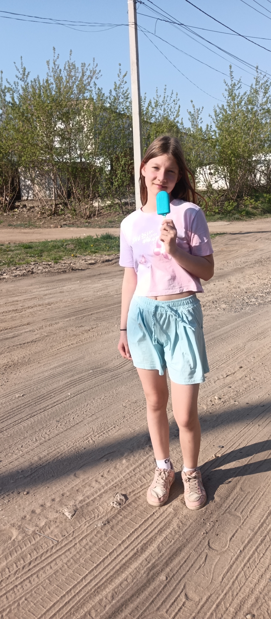 A full-length shot reveals a young woman standing on a dirt road in Unknown, Unknown. She holds a blue popsicle to her mouth with her right hand. The subject wears a light-purple and pink tie-dye crop-top shirt, light blue shorts with a drawstring, white socks, and dusty pink sneakers. A black bracelet adorns her left wrist. The dirt road shows tire tracks and scattered debris. Behind her, bushes and trees are visible alongside a white utility pole. The sky is a clear blue.