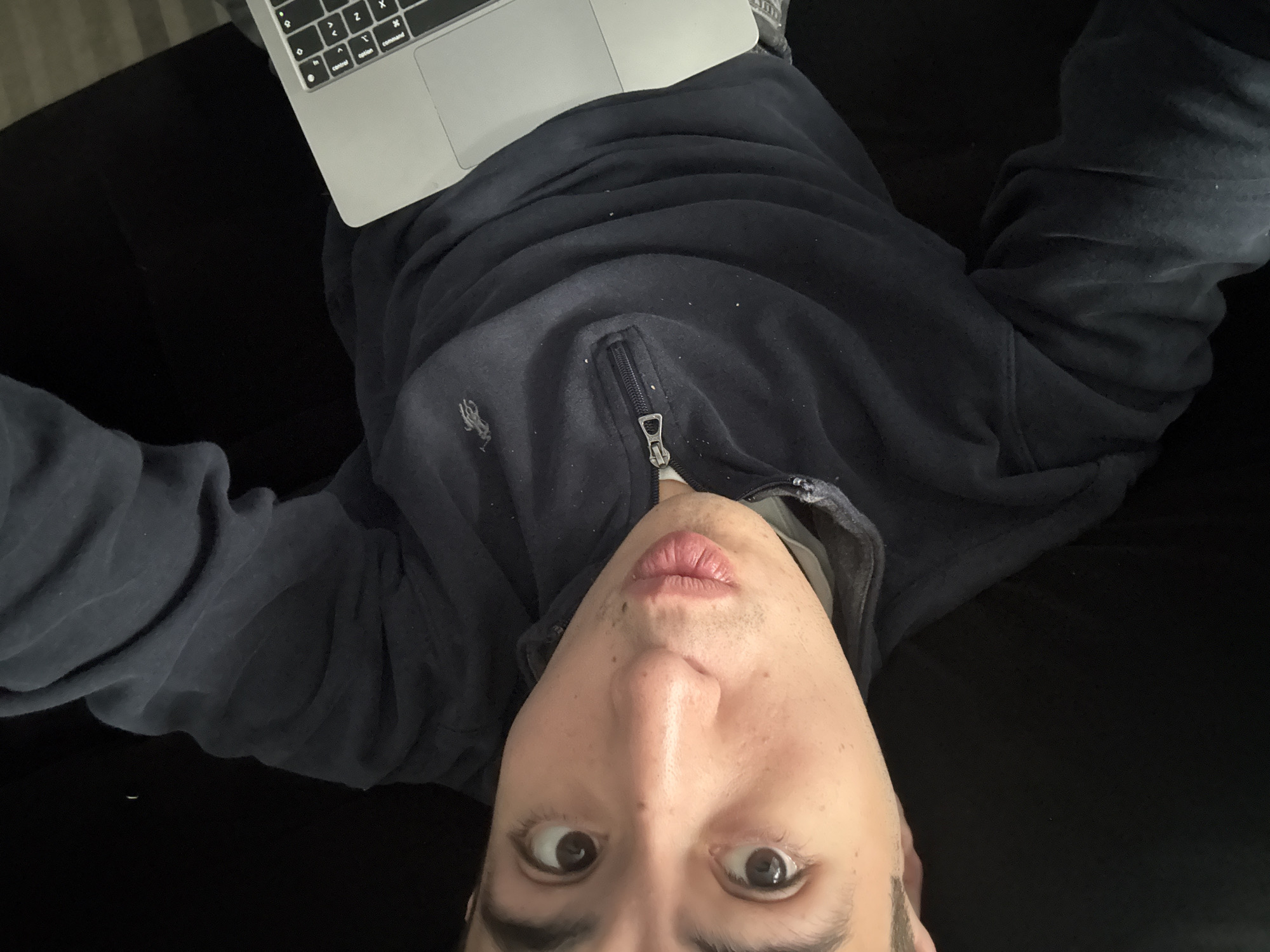 A person is lying on a black couch, looking upwards into the camera. They are wearing a dark blue zipped sweatshirt and have a laptop resting on their torso. The location is not identifiable as San Bernardino Tlaxcalancingo, Mexico based on the visible content.