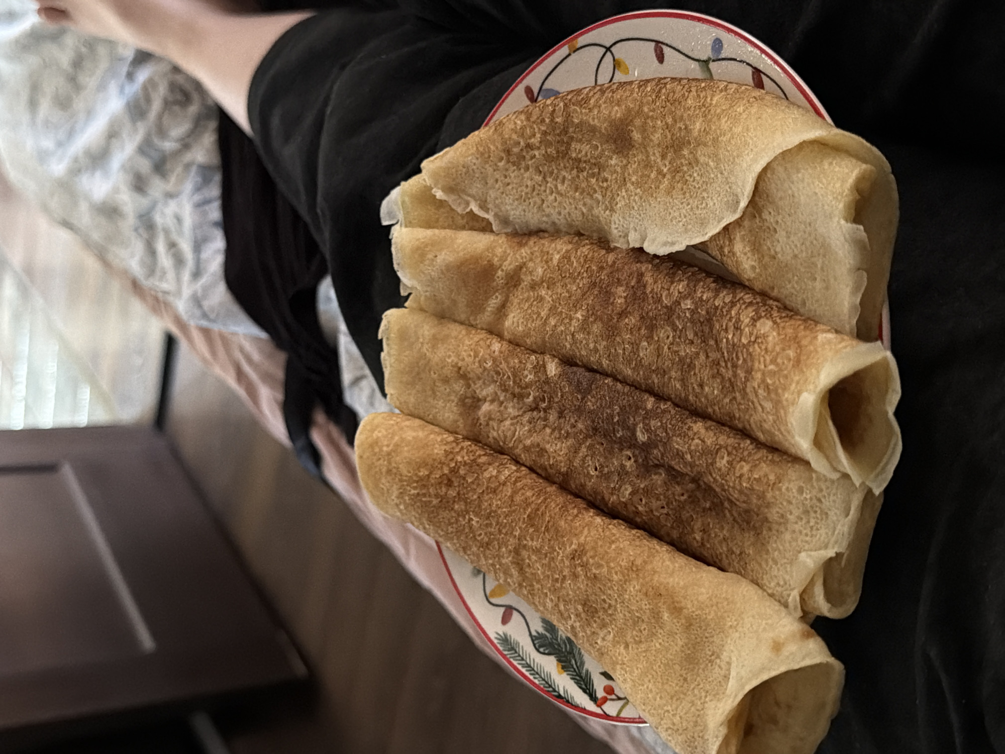 In a close-up shot, four rolled crepes are presented on a plate with a festive holiday design of colorful lights and berries. The crepes are golden brown with slightly darker browning in some areas, suggesting they have been cooked to a crisp. They are arranged diagonally on the plate, with the open ends facing the viewer. The plate rests on a dark, soft surface, possibly clothing or bedding. In the background, a sliver of a dark brown wooden door is visible to the left, and a blurred expanse of white and pink patterned fabric occupies the upper right, hinting at a bed. A person's legs, partially visible on the right side of the frame, are clad in black shorts or pants, and their skin is lightly tanned. The lighting suggests an indoor setting, possibly a home, with natural light filtering in from a window out of frame. The scene is devoid of any specific landmarks or features that would indicate a location in Tbilisi, Georgia.