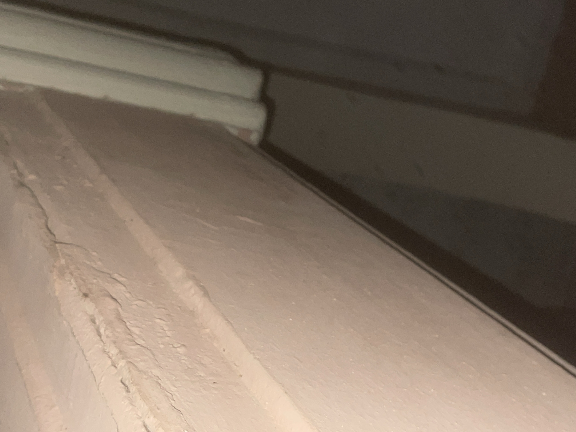 The image shows a close-up view of a painted wooden surface, possibly a window sill or trim, with peeling and cracked paint. The surface is a light pink or beige color and shows signs of age and wear. To the right, a vertical white object, likely a door frame or part of a wall, is visible. The background is dark and out of focus, suggesting an indoor environment. The scene is located in Howayi, Nigeria. There are no people or visible actions occurring in the image.