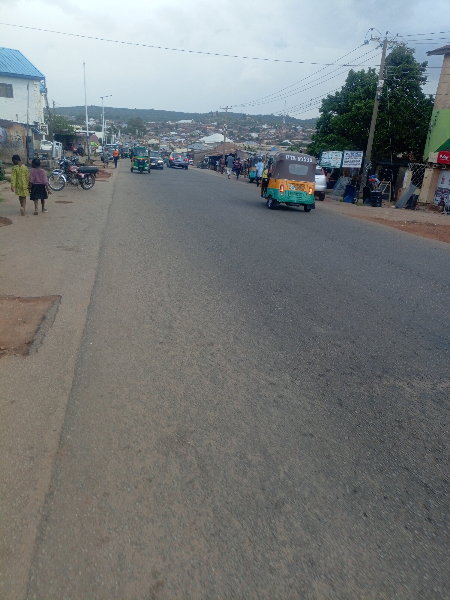 The image depicts a street scene in Jos, Nigeria. A paved road occupies the foreground and midground, with a slight upward slope towards the horizon. On the left side of the road, a sidewalk is visible, bordered by a dirt and gravel area. Two young girls are walking on the sidewalk, one wearing a yellow dress and the other a pink top and dark skirt. A motorcycle is parked on the sidewalk behind them.
Further down the road, several vehicles are in motion and parked. A green and yellow auto-rickshaw, identified by the license plate PTA-10591, is moving away from the camera. Other cars and vehicles are also present in the street. The roadside on the right is lined with buildings and businesses, some with awnings and signage. A prominent green structure with a red "Palrnet" sign is visible. Power lines and utility poles are strung across the scene, with trees and hills forming the background, indicative of the hilly terrain of Jos. The sky is overcast. The scene appears to be a typical urban street with moderate activity.