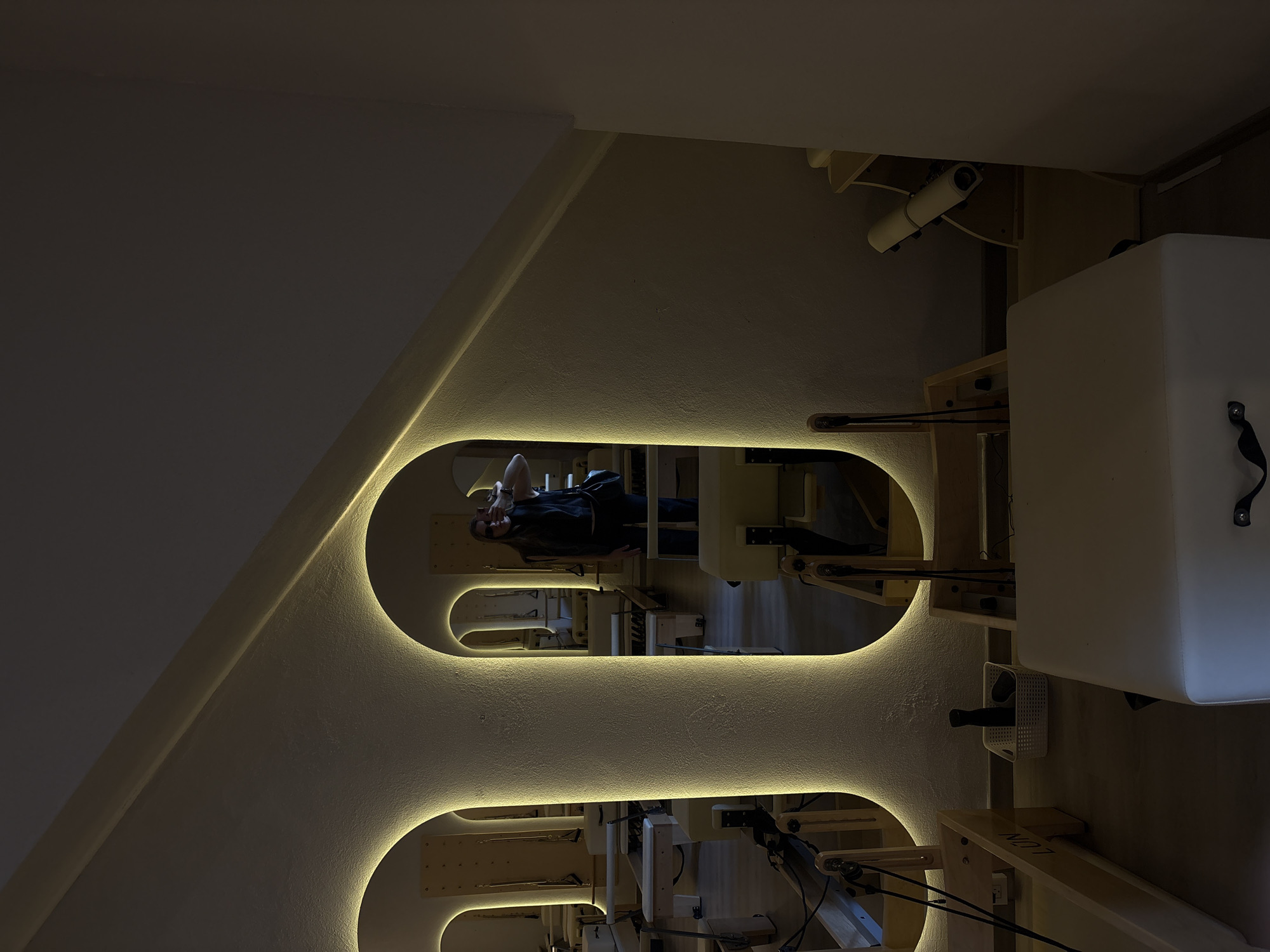 The image shows the interior of a pilates studio, likely in Mexico City, Mexico, based on the presence of a person reflected in a rounded wall-mounted mirror. The mirror is illuminated with warm, glowing lights that accentuates its unique shape, resembling a spine or an abstract art form. The reflection shows a person lying on a pilates reformer machine, possibly exercising or preparing for a session. Various pilates machines are arranged around the room. The room has a neutral color palette, with the white walls, beige toned pilates machines, and warm light providing a calm and inviting atmosphere for exercise and wellness activities.