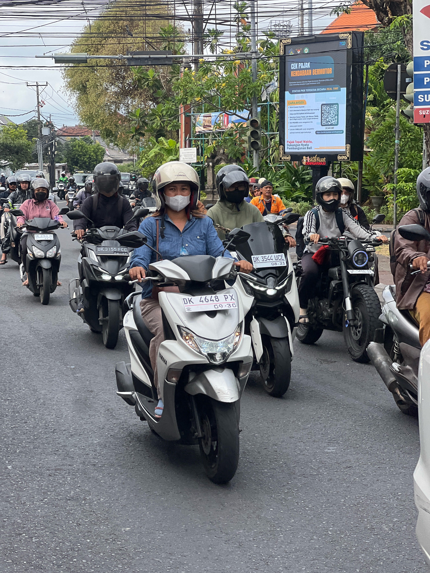 This is a medium-shot, eye-level, outdoor photograph taken from behind a motorcyclist, looking forward at a street scene in Denpasar, Indonesia. The image depicts a busy street with multiple motorcycles and riders, indicative of urban traffic congestion. The riders, predominantly wearing helmets and some with face masks, are navigating the paved road. Several motorcycles are clearly visible, with license plates such as "DK 3743 AI", "DK 3155 FCC", and "DK 4648 PX" identifiable. A prominent digital billboard on the right side of the frame displays information about checking motorcycle tax in Denpasar. The environment includes overhead power lines, lush green trees, buildings, and traffic lights, suggesting a typical urban street in a tropical climate. The overall atmosphere is one of daily commuting and the hustle and bustle of city life.