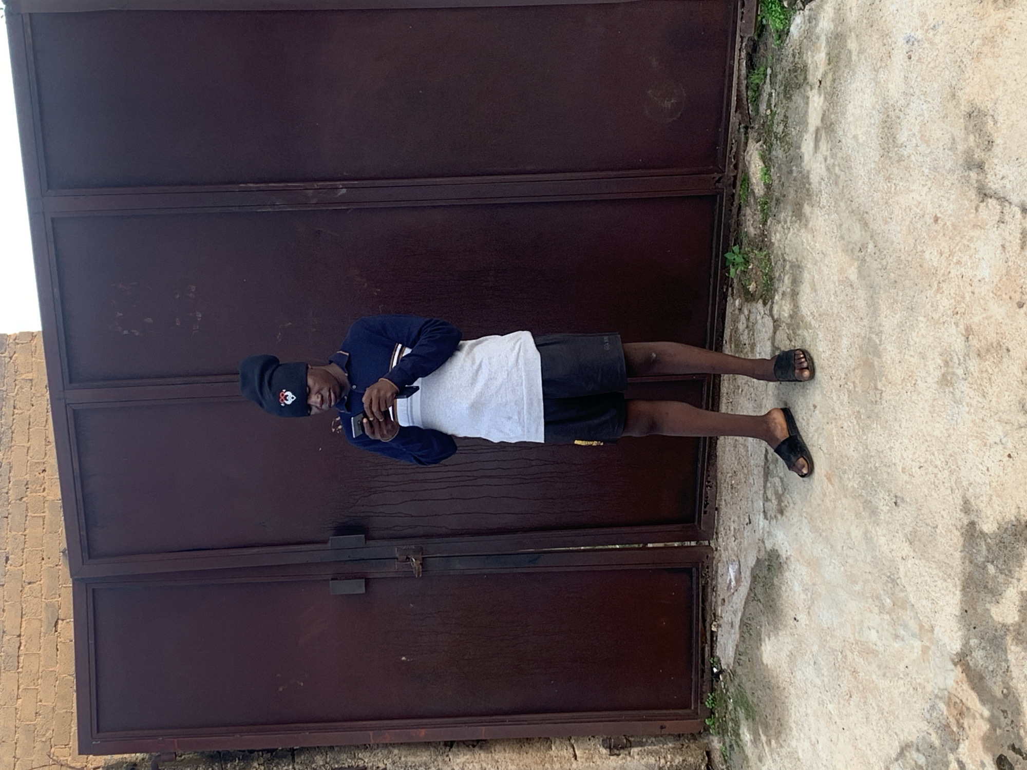 A young Black man is standing in front of a large, dark brown metal double door with a brick wall above it. The man is wearing a black beanie with a red and white logo, a dark blue long-sleeved shirt with a white horizontal stripe across the chest, and black shorts. He is holding a smartphone in his hands and appears to be looking down at it. He is wearing black slides on his feet. The ground in front of the doors is concrete with some patches of dirt and grass. The overall scene suggests an outdoor urban or semi-urban environment in Gura Top, Nigeria.