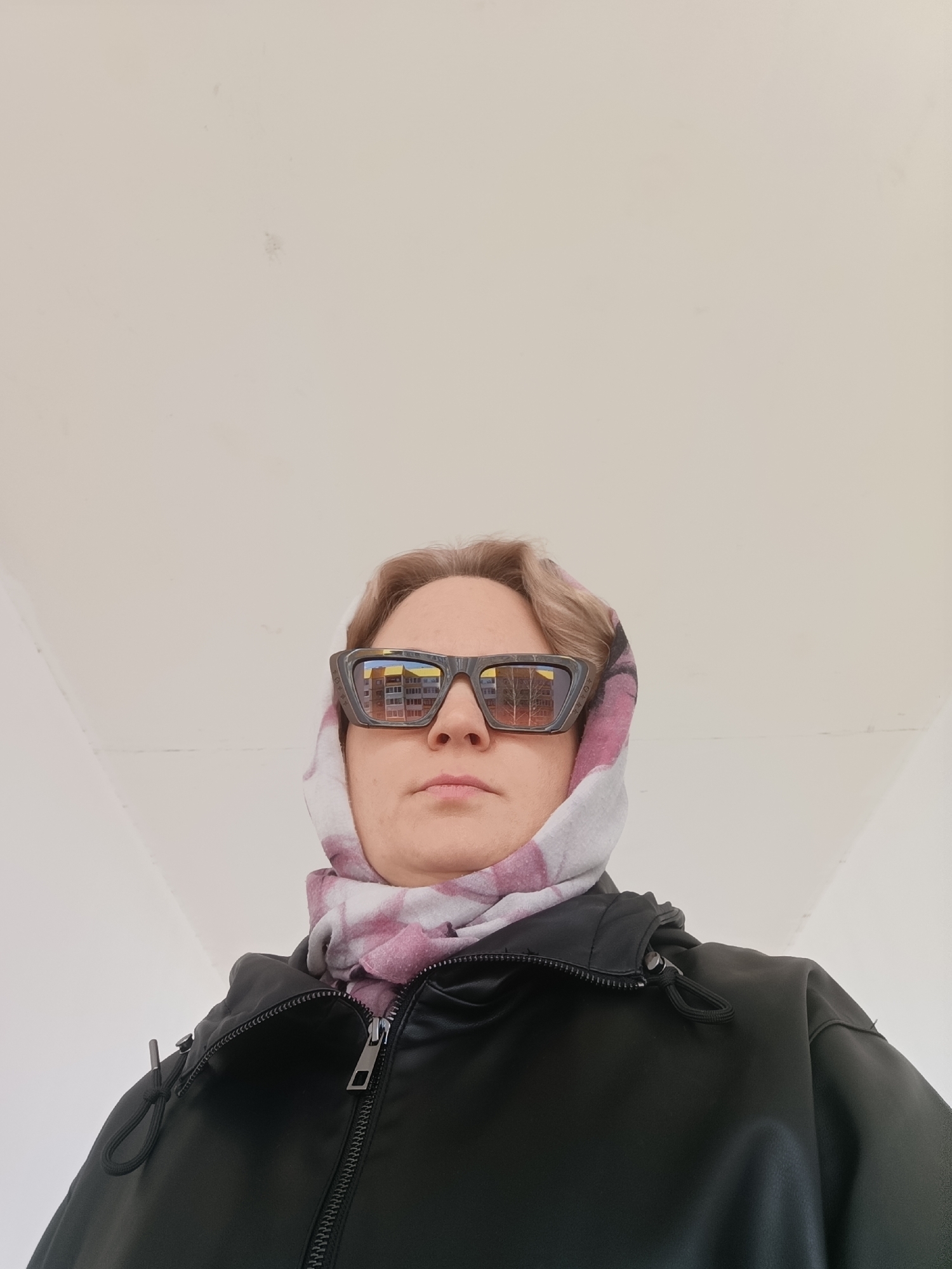 A person wearing a black jacket and a headscarf stands indoors, viewed from a low angle, with a plain white ceiling in the background. The headscarf has a pale, checkered pattern with pinkish-red hues. The person is also wearing dark-framed sunglasses that reflect a building with orange and grey-brown tones. The person's facial expression is neutral. Based on the reflection in the sunglasses, the location appears to be in Марковский, Russia.
