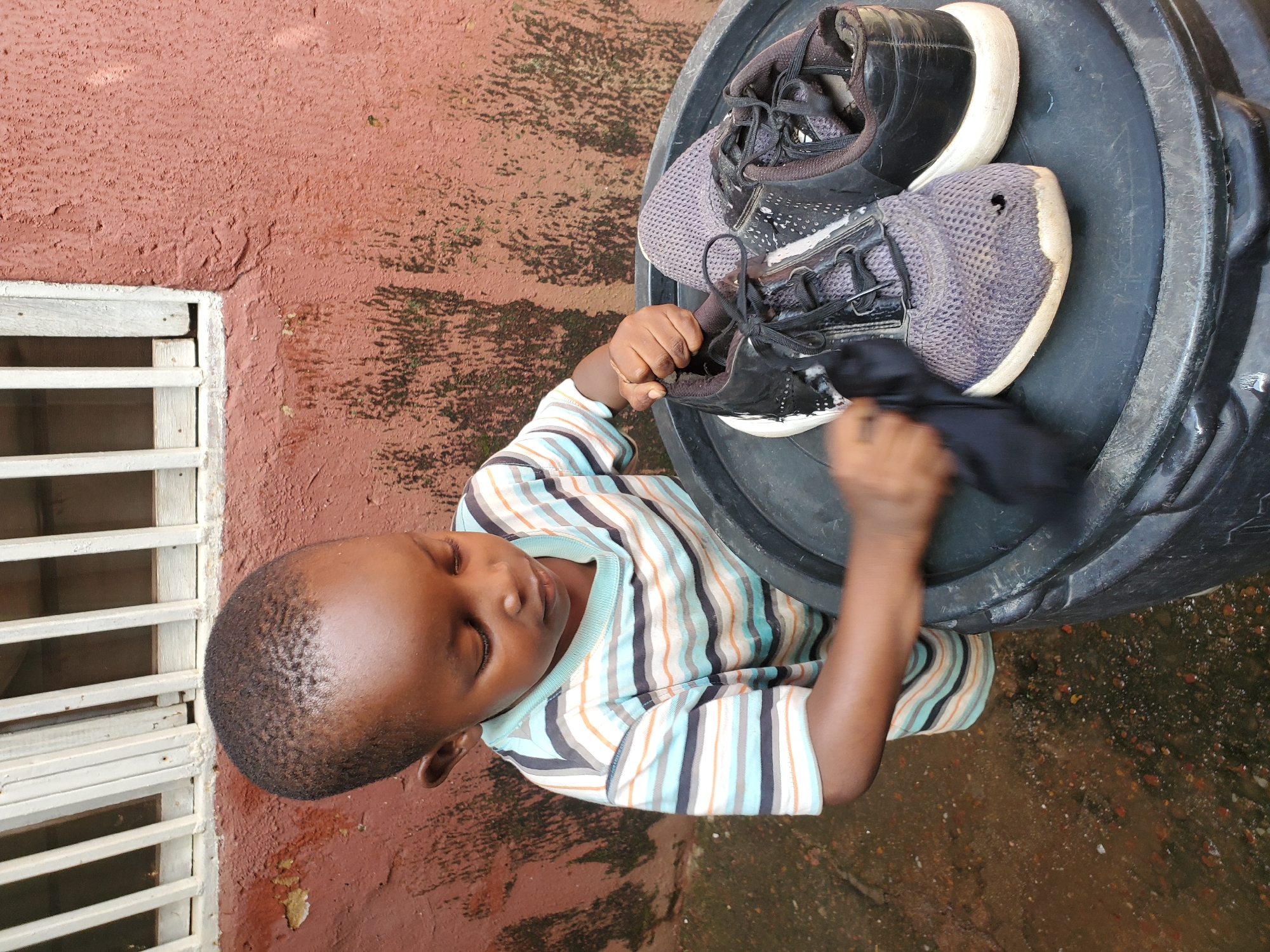 The image shows a young child, likely a boy, wearing a striped t-shirt in shades of blue, orange, and white. He is sitting on what appears to be a large black barrel, holding a black cloth and a pair of dark-colored athletic shoes. The shoes are partially wet and appear to be mesh with white soles and black laces. The child is looking down at the shoes, seemingly engaged in cleaning or examining them. His eyes are closed, and his expression suggests concentration or perhaps mild effort.
The setting appears to be outdoors, with a reddish-brown, textured wall visible to the left, featuring a white-barred window or grill. The ground is a mix of reddish-brown earth and scattered stones, with some patches of wetness, indicating it might be damp or has recently rained. The overall lighting suggests it could be daytime, possibly under an overcast sky given the diffused light. There are no visible signs or text in the image. The scene conveys a sense of domestic activity, possibly a chore or play involving the shoes and the cloth. The location context of Jos, Nigeria, suggests a warm climate and potentially a casual, everyday environment.