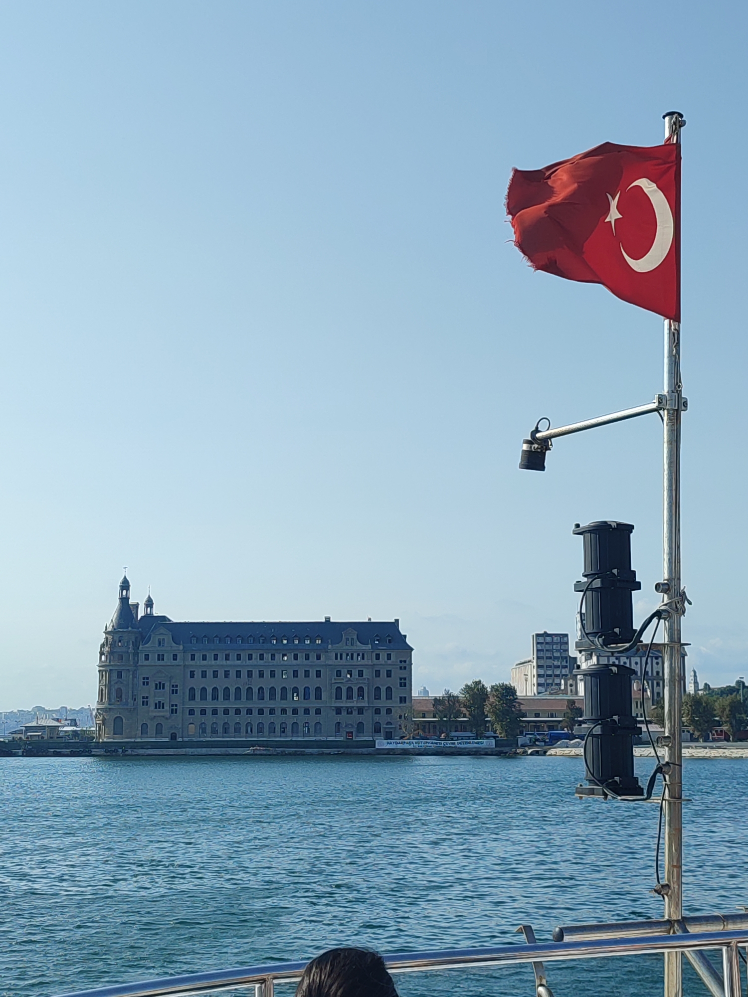 The image depicts a view from a boat on the water, looking towards the Kadıköy district of Istanbul, Turkey. In the foreground, the edge of a metal railing is visible, with a person's hair at the bottom of the frame. A tall flagpole stands to the right, flying a Turkish flag that is slightly torn at the edges. Attached to the flagpole are two black, cylindrical objects, possibly speakers or lights, and a security camera.
In the middle ground, across the water, a large, historic building with numerous windows and two distinctive towers is prominently featured. This building is identified as the Haydarpaşa Train Station. Behind the station, other buildings and trees are visible, suggesting a city skyline. The water is a deep blue, with ripples indicating movement. The sky is clear and pale blue. The overall scene captures a waterfront view of Kadıköy with its iconic train station.