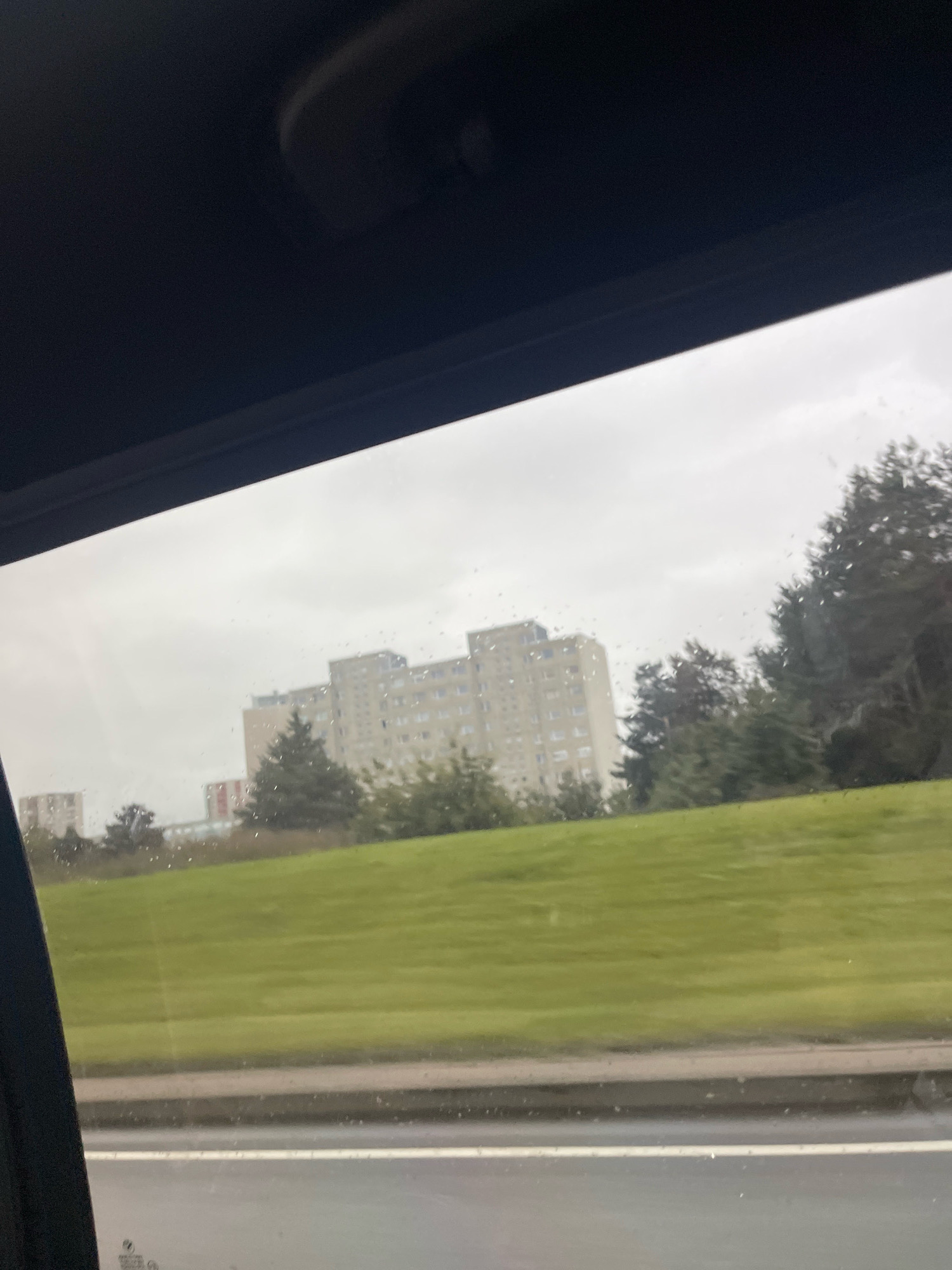 The image captures a view through a car window, looking out at a cityscape under an overcast sky. In the background, several tall, light-colored apartment buildings are visible, with one prominent building appearing lighter than the others, suggesting it might be made of concrete. A green, grassy hill slopes upwards towards the buildings. There are trees scattered across the landscape, including some evergreen trees that are darker than the deciduous trees. The foreground shows a road with a white line. The window itself has water droplets on it, indicating it might be raining. The overall impression is that of a suburban or residential area in Tallinn, Estonia, viewed from a moving vehicle.