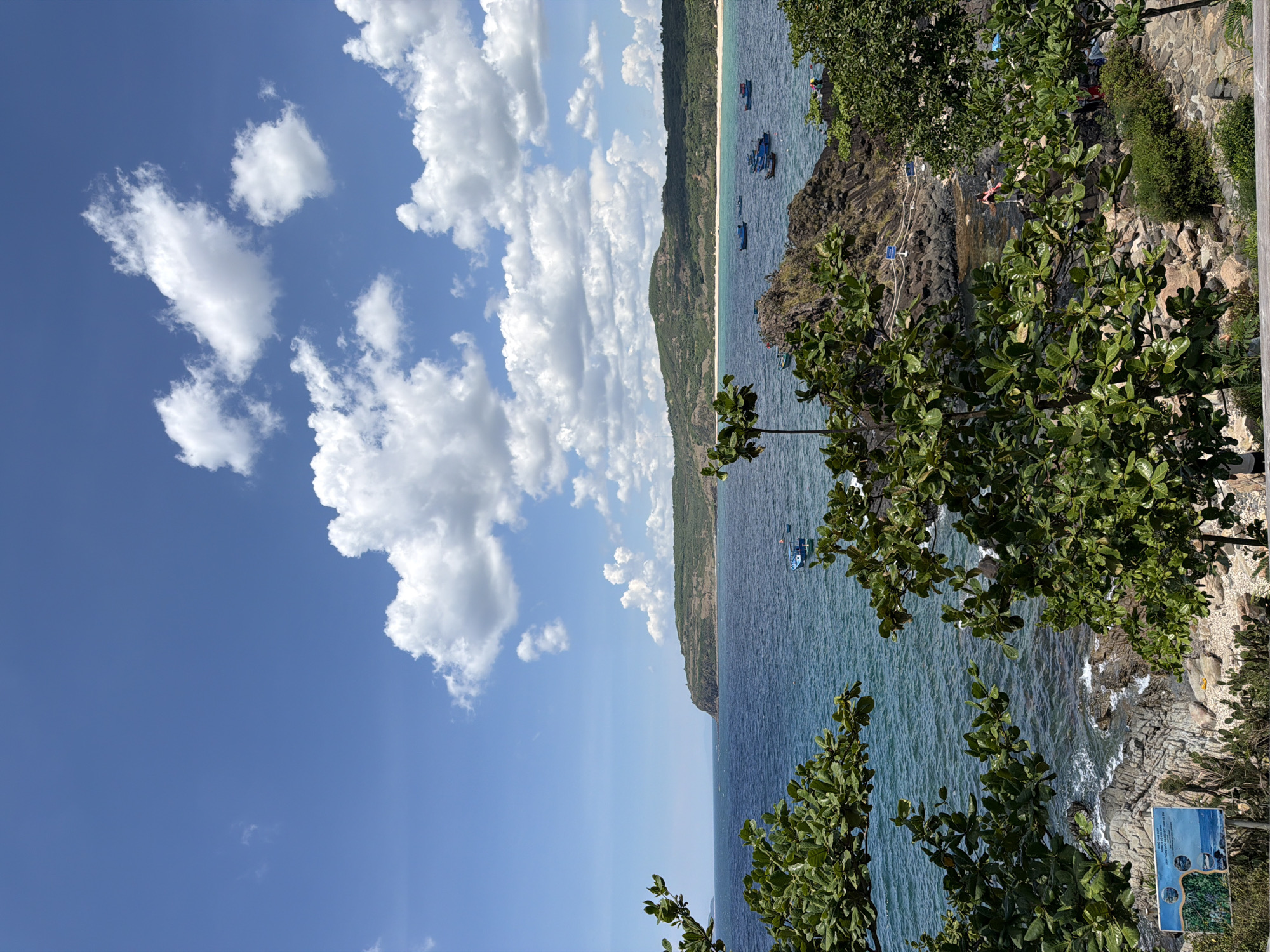 The image depicts a picturesque coastal scene in Phú Lương, Vietnam, under a bright, partly cloudy blue sky. In the foreground, lush green trees with broad leaves frame the view. Beyond the trees lies a rocky shoreline and the calm, blue sea. Several small, blue fishing boats are dotted across the water, some closer to the shore and others further out. In the mid-ground, a sandy beach stretches along the coast, backed by rolling hills covered in dense green vegetation. The distant landscape is characterized by these verdant hills under the expansive sky. A few people are visible near the rocky areas along the shore.
