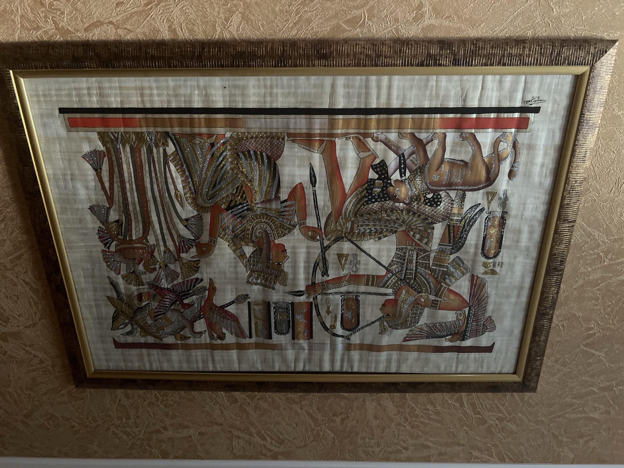 This image shows a framed papyrus painting depicting an ancient Egyptian scene. The painting is hung on a textured, light brown wall. The artwork features two central figures, one appearing to be a pharaoh drawing a bow and arrow, and another seated figure with a headdress. A lion is positioned to the left of the pharaoh, and a collection of birds is depicted on the right. Hieroglyphics and other Egyptian motifs are visible throughout the painting. The frame is ornate, with a golden inner border and a darker outer frame with a textured finish. There is no discernible evidence in the image to indicate a specific location within the city of Tula, Russia.