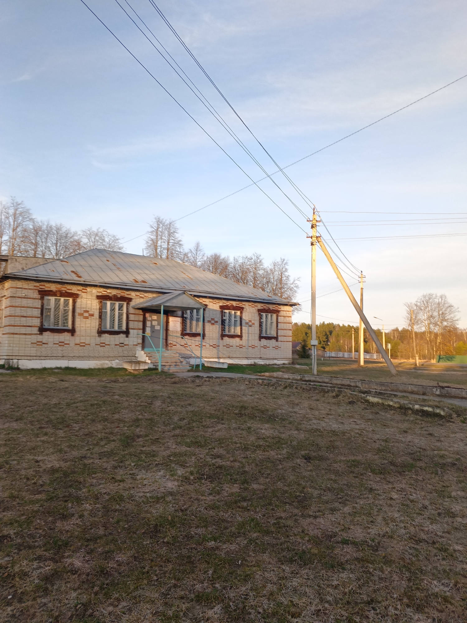The image shows a single-story brick building with a metal roof in Красный Маяк, Russia. The building has several windows with dark brown trim. There is a small porch with a roof supported by light blue metal poles. The ground in front of the building is covered in brown grass. Power lines stretch across the sky, connected to a wooden utility pole with a support arm. In the background, trees and another utility pole are visible. The sky is light blue with some white clouds.