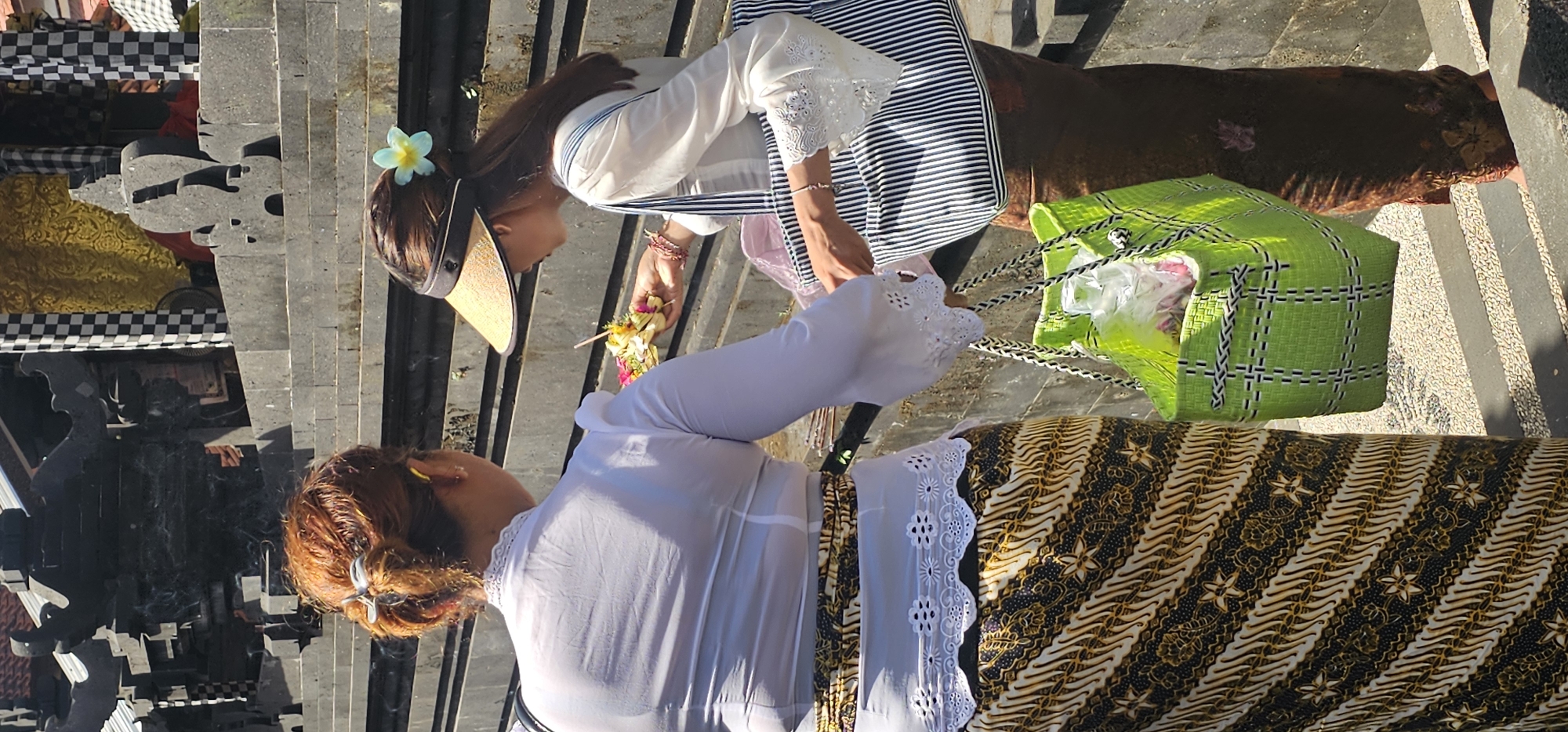 Two women are on a stone staircase, possibly at a temple in Kuta, Indonesia. Both are dressed in traditional Balinese attire. The woman on the left has reddish-brown hair, a white blouse with lace trim, and a batik sarong. The woman on the right wears a sun visor and a flower in her hair, a white blouse with lace, and a striped sarong. She carries a green woven bag. Both are placing small floral offerings on the stairs. To the left, one can see the stone carvings of the temple, adorned with checkered cloths and gold fabrics. The setting appears sunny.
