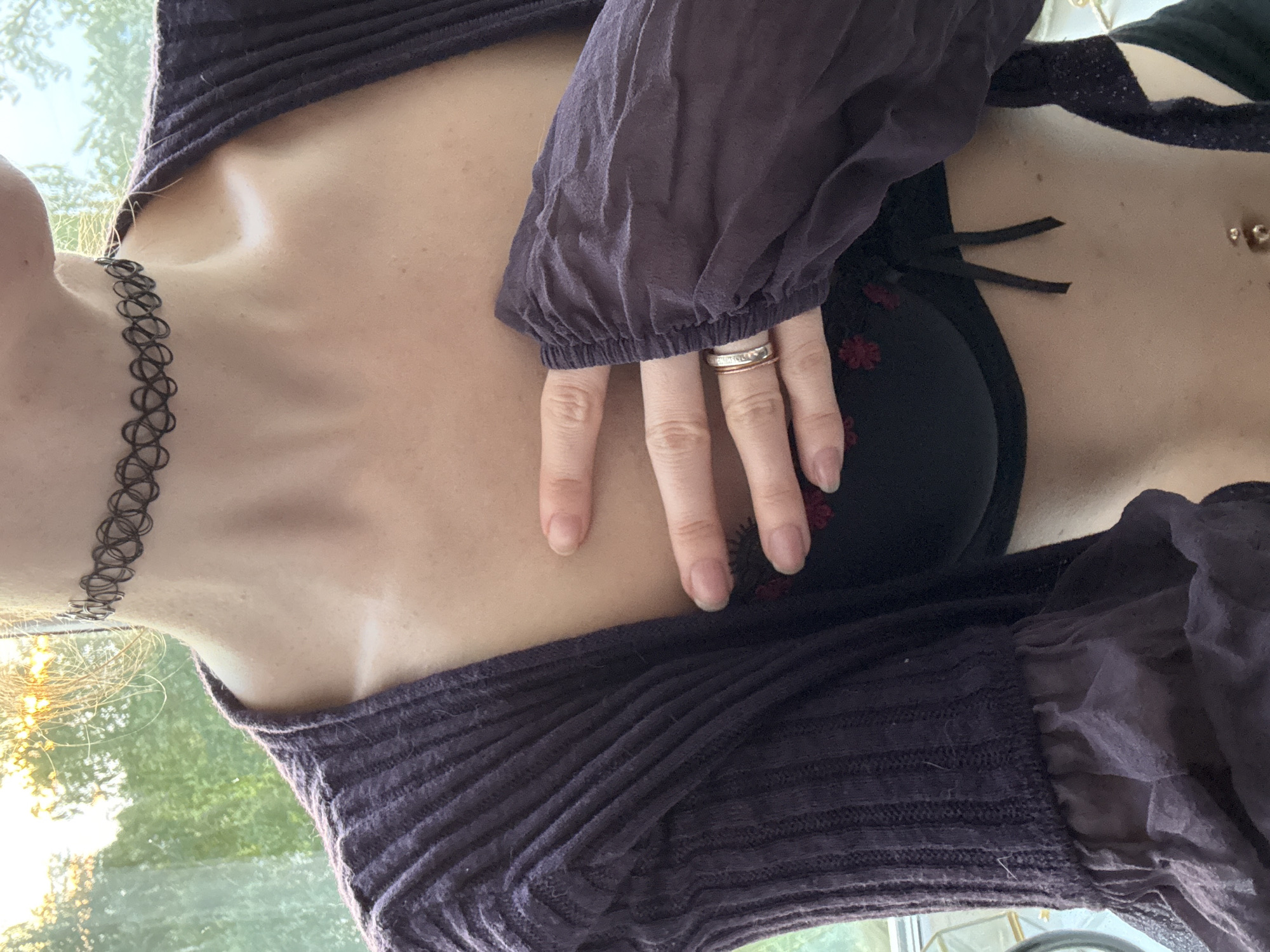 A close-up, chest-level shot focuses on a person wearing a dark purple cardigan over a black bra with red floral embroidery. The cardigan has ribbed sleeves with ruffled cuffs made of sheer fabric. The person is wearing a black choker necklace and two rings on their right hand, one of which appears to be a thin rose gold band. Their right hand is placed over their chest. A belly button piercing is visible. The background is blurred, showing a window with trees and sunlight filtering through. The setting does not provide specific details to indicate a location in Chaikovsky, Russia.