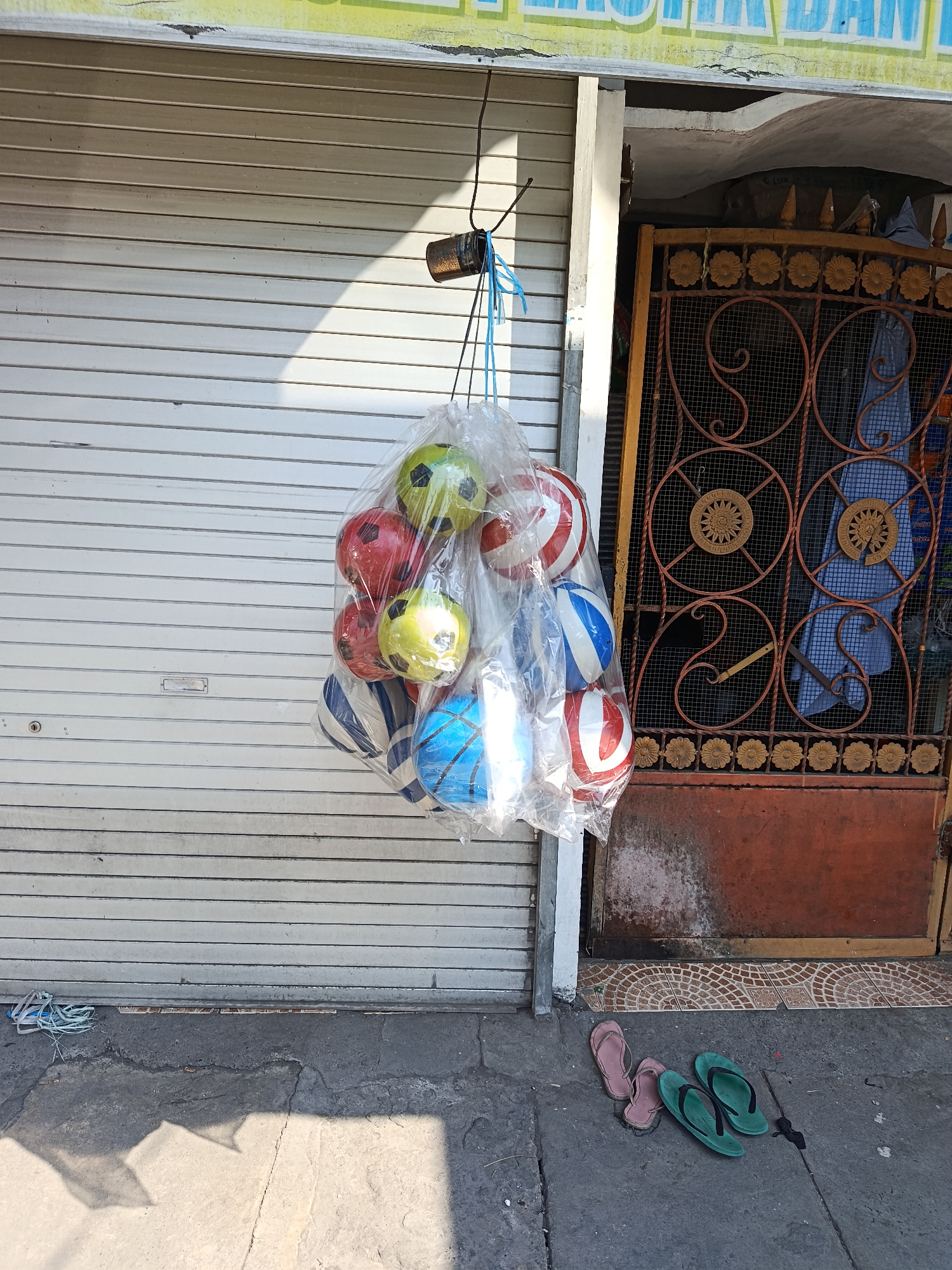 In Keboansikep, Indonesia, a bundle of colorful balls, encased in clear plastic, hangs from a hook on a weathered metal security gate. The balls, in shades of red, yellow, blue, and white with black accents, appear to be children's toys. They are suspended in front of a rolled-up metal storefront, its corrugated surface reflecting the bright sunlight. The gate itself is adorned with intricate floral patterns and circular motifs in rust-colored metal, providing a stark contrast to the plain storefront. To the right of the gate, a blue garment is visible. On the ground in front of the storefront, a pair of pink flip-flops and two green flip-flops are casually placed, suggesting recent human presence. The pavement is cracked and shows signs of wear, with a shadow cast from an unseen object. The overall scene depicts a common street-side retail display in an urban Indonesian setting.