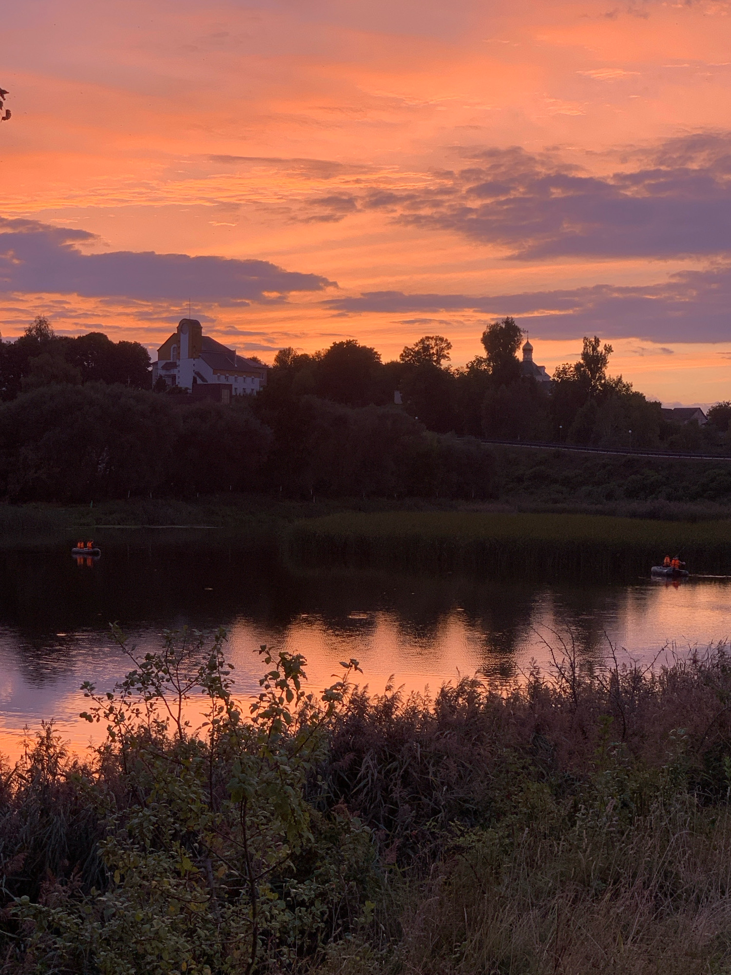 An outdoor, eye-level shot captures a body of water at dusk, with a vibrant sunset painting the sky in hues of orange and purple. Silhouetted trees line the distant shore, punctuated by the architectural features of a church and a domed building. Two small boats with people wearing orange life vests are visible on the water. The foreground is filled with dense, dry vegetation. The scene appears to be the Pripyat River in Kapyĺ, Belarus.